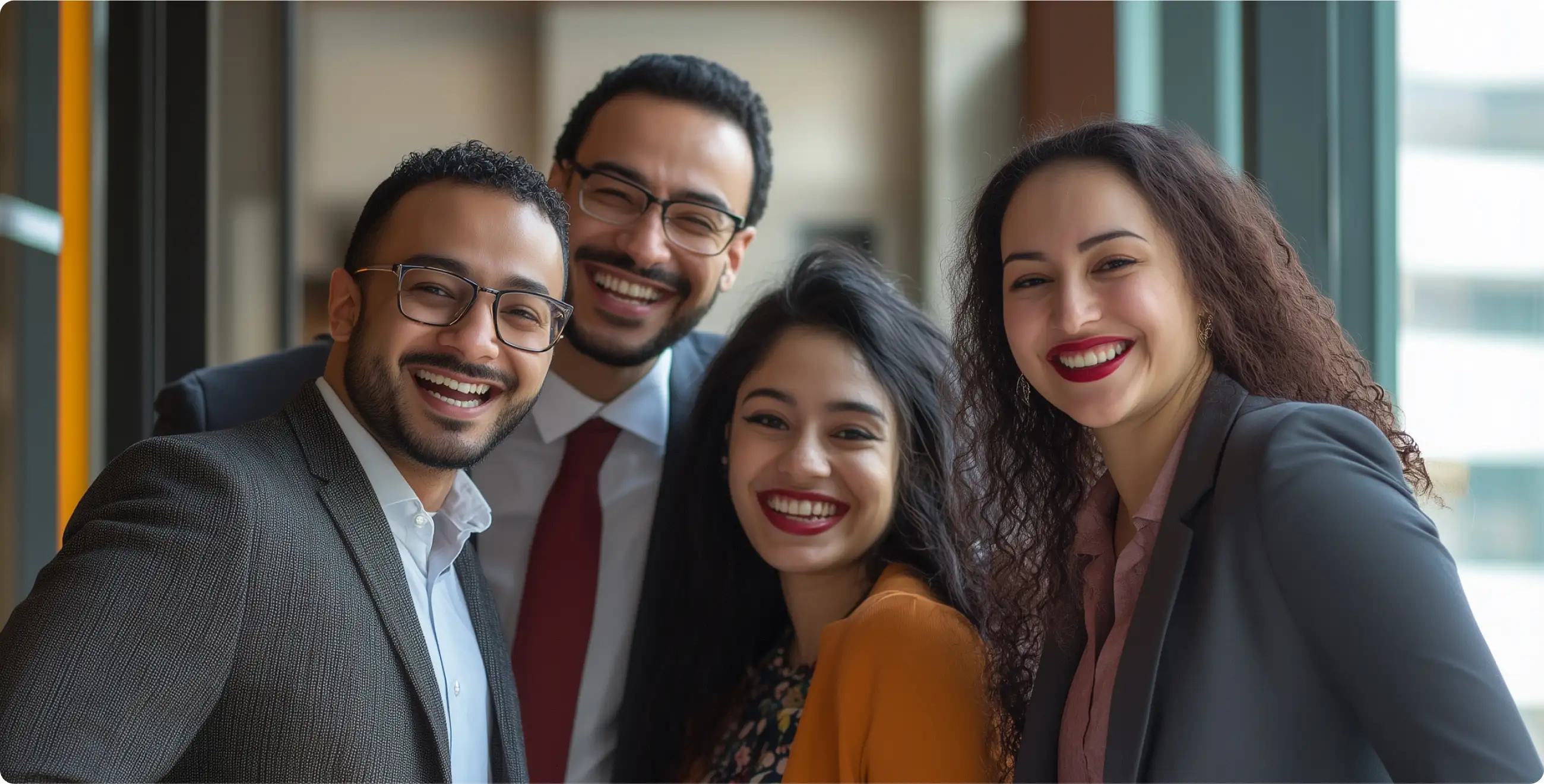 A group of four individuals stands together, dressed in professional attire, posing near large windows with a modern office backdrop.