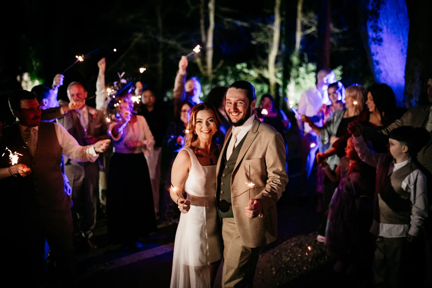 Smiling bride and groom holding sparklers surrounded by guests with sparklers at nighttime outdoor wedding celebration.