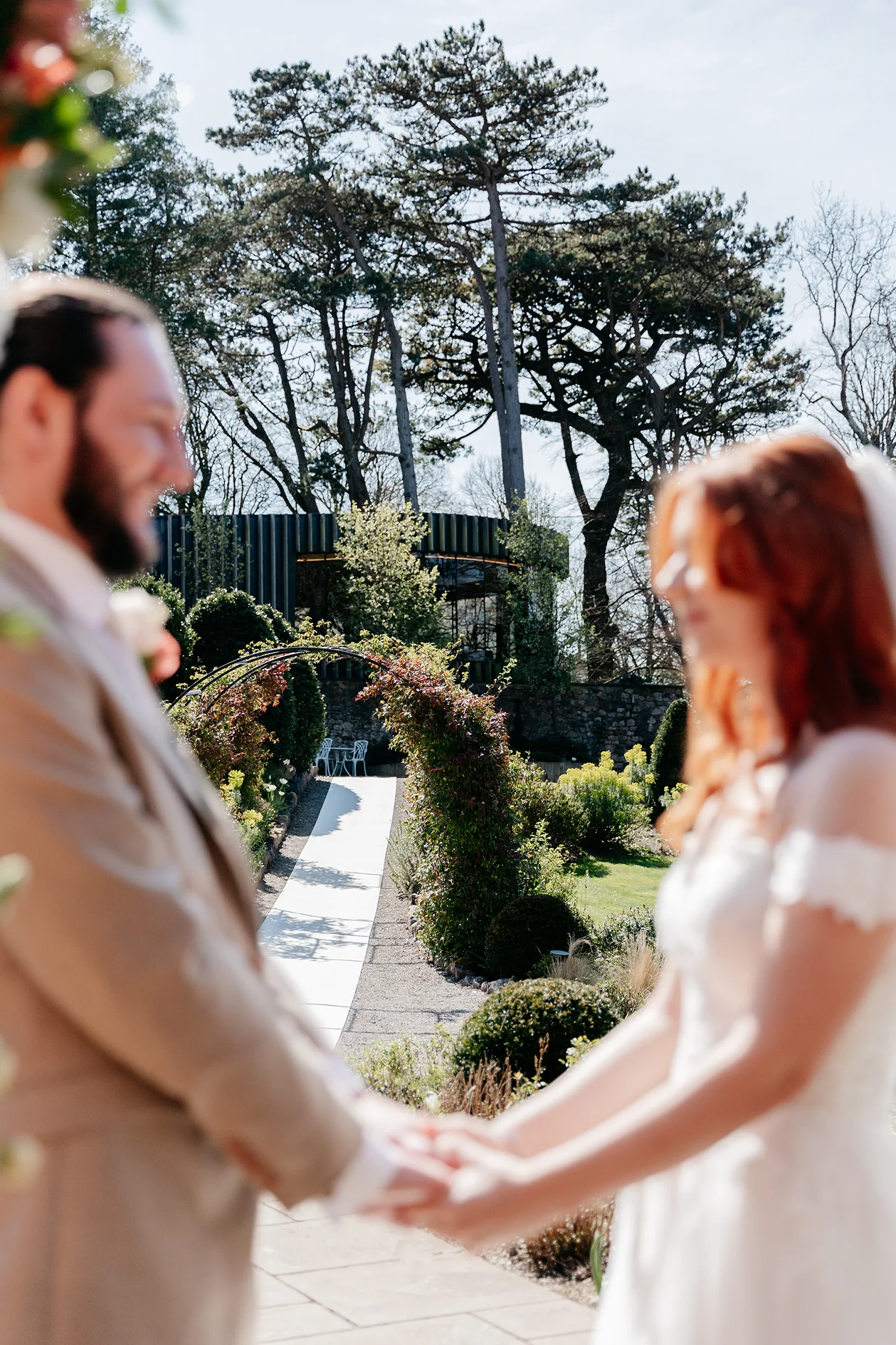 Blurred image of a bride and groom holding hands outdoors with a garden path and trees in the background.