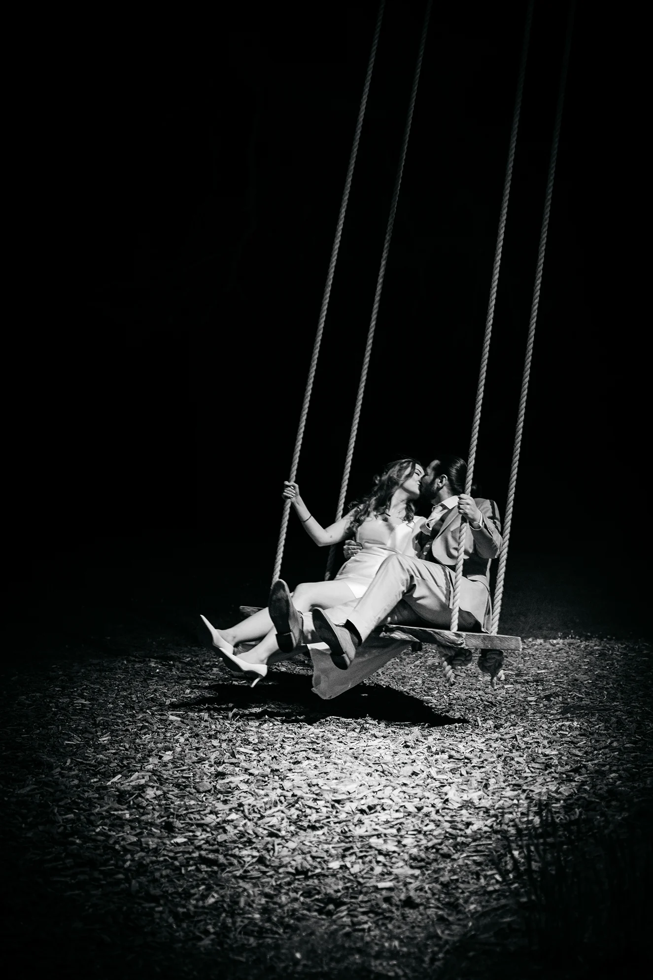 Black and white photo of a couple dressed formally kissing while sitting together on a swing at night.