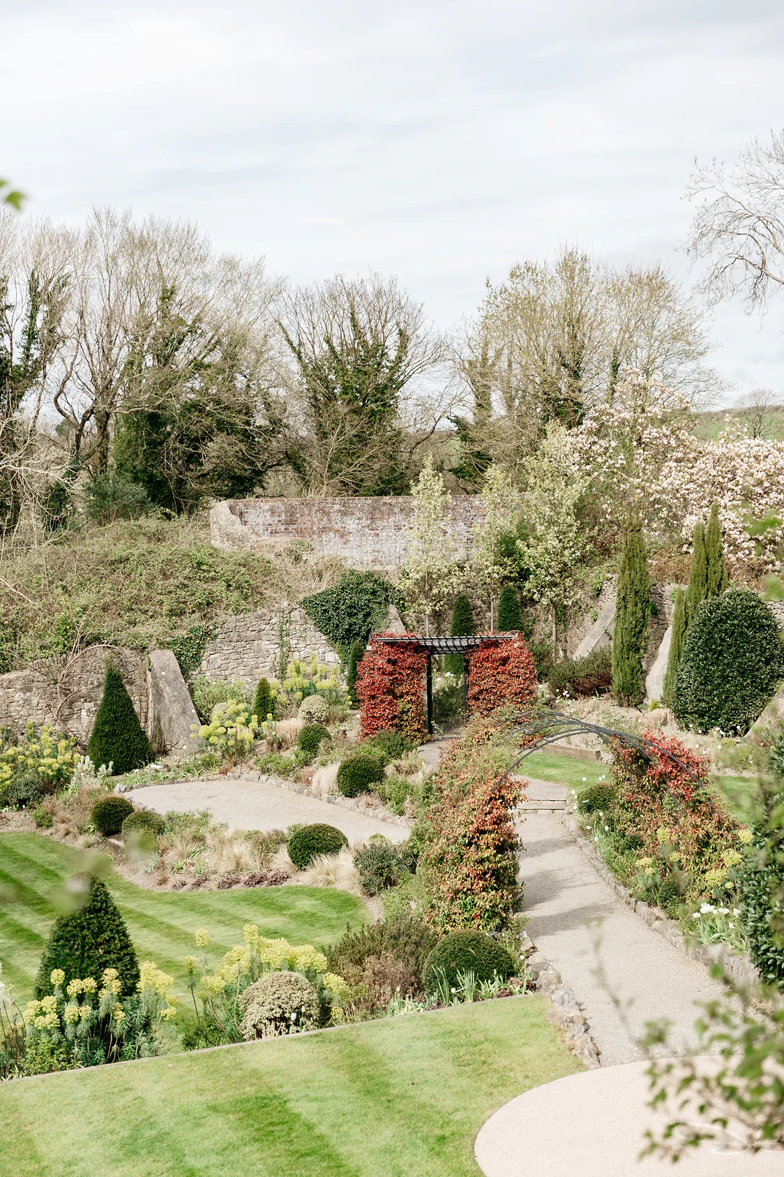 Ornamental garden with trimmed bushes, climbing plants on arches and a stone wall in the background.
