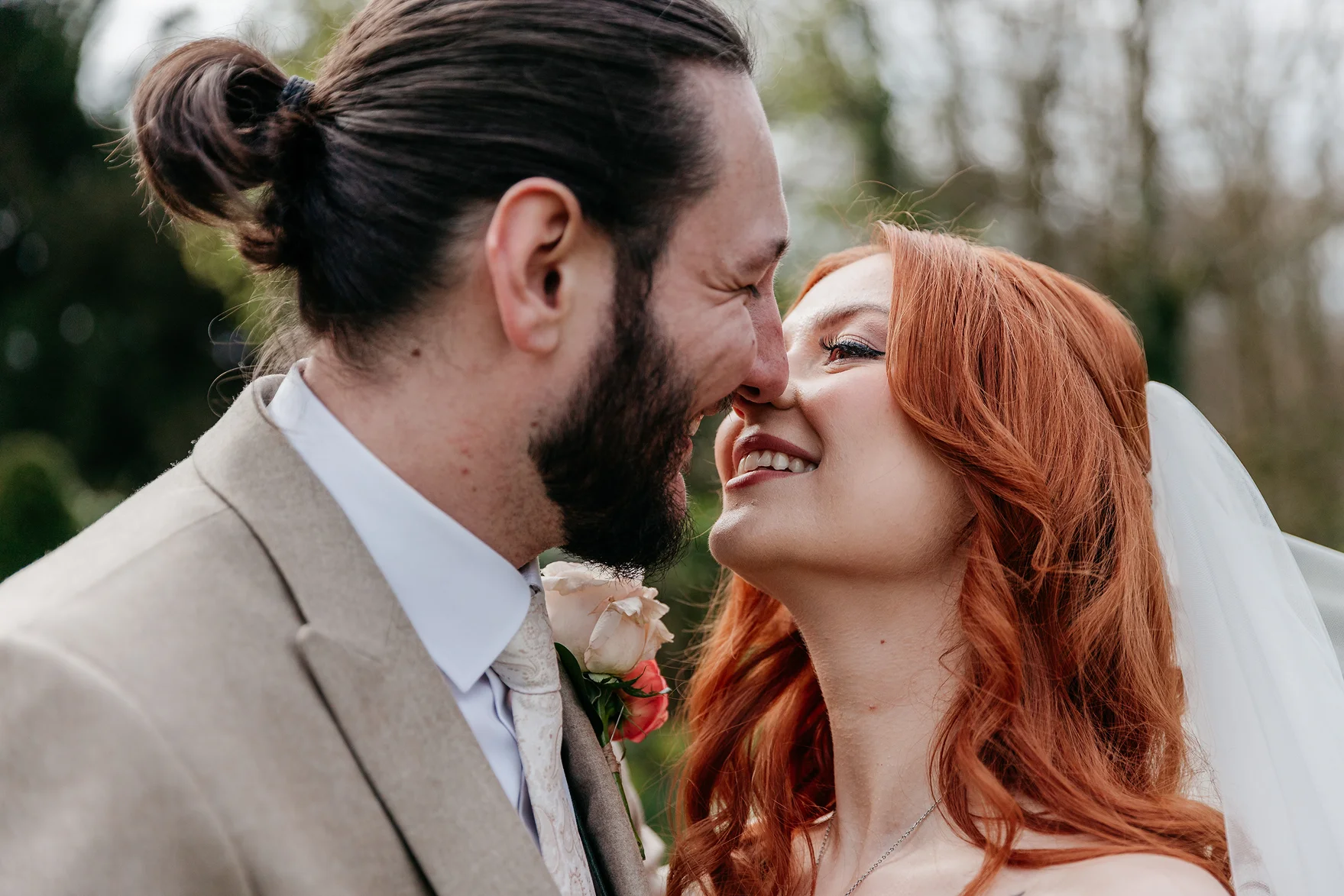 Close-up of a smiling bride with red hair and a veil touching noses with a groom in a beige suit during their wedding.