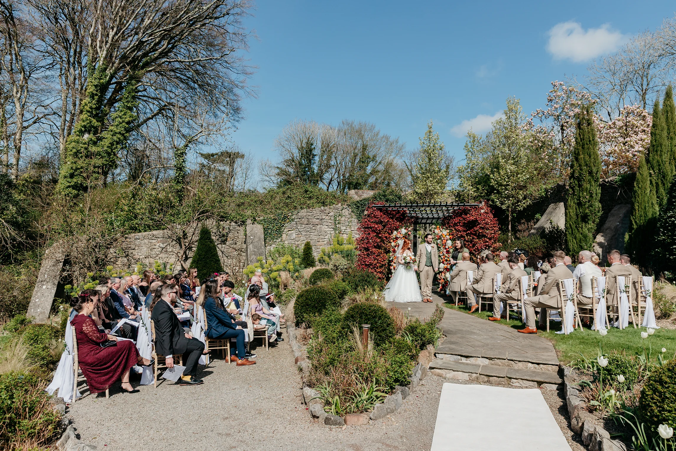 Outdoor wedding ceremony with bride and groom standing under a floral arch surrounded by guests seated on both sides.