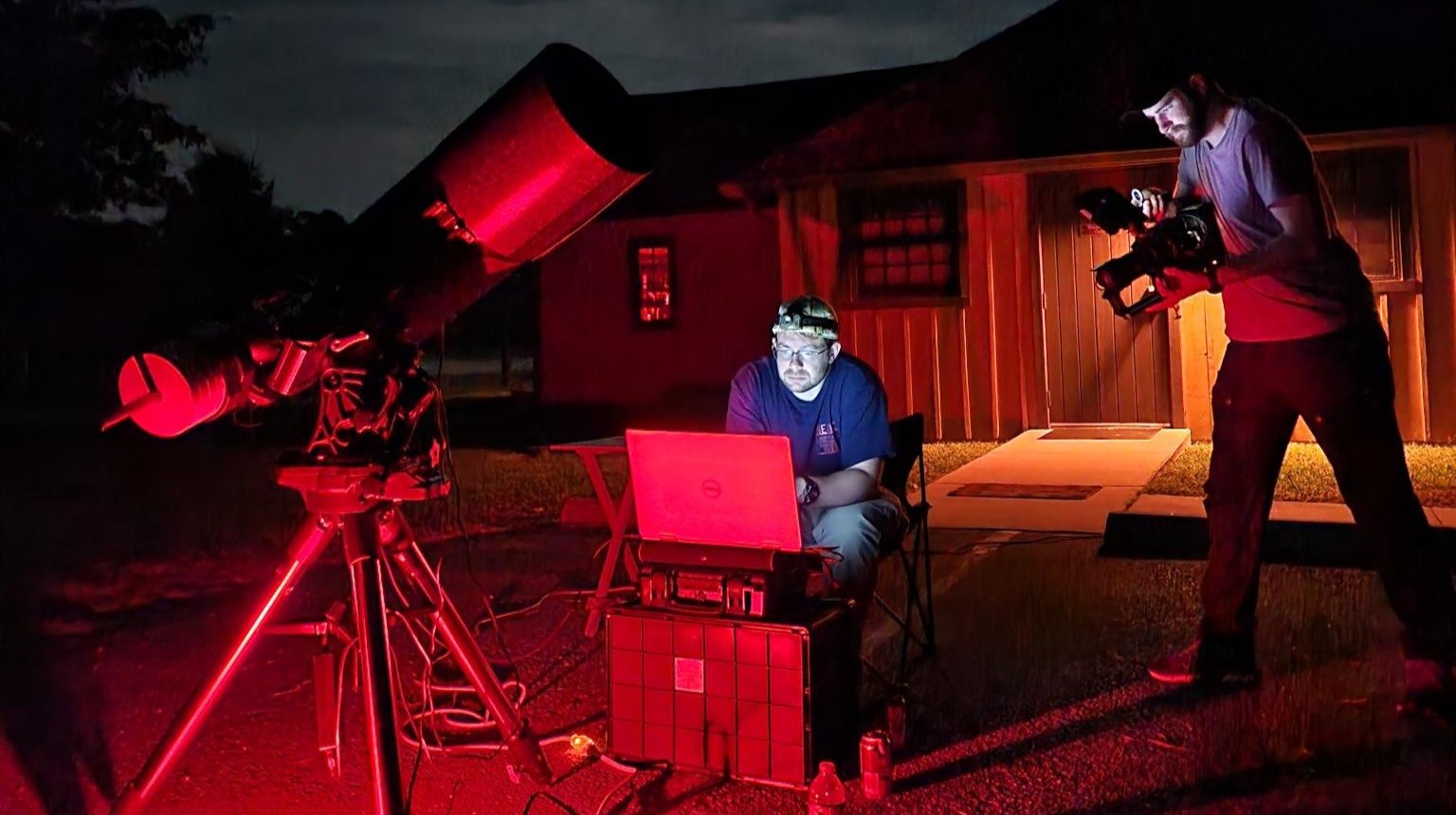 Two men at night with a large telescope and a laptop, one operating the laptop illuminated by red light, the other filming with a video camera.