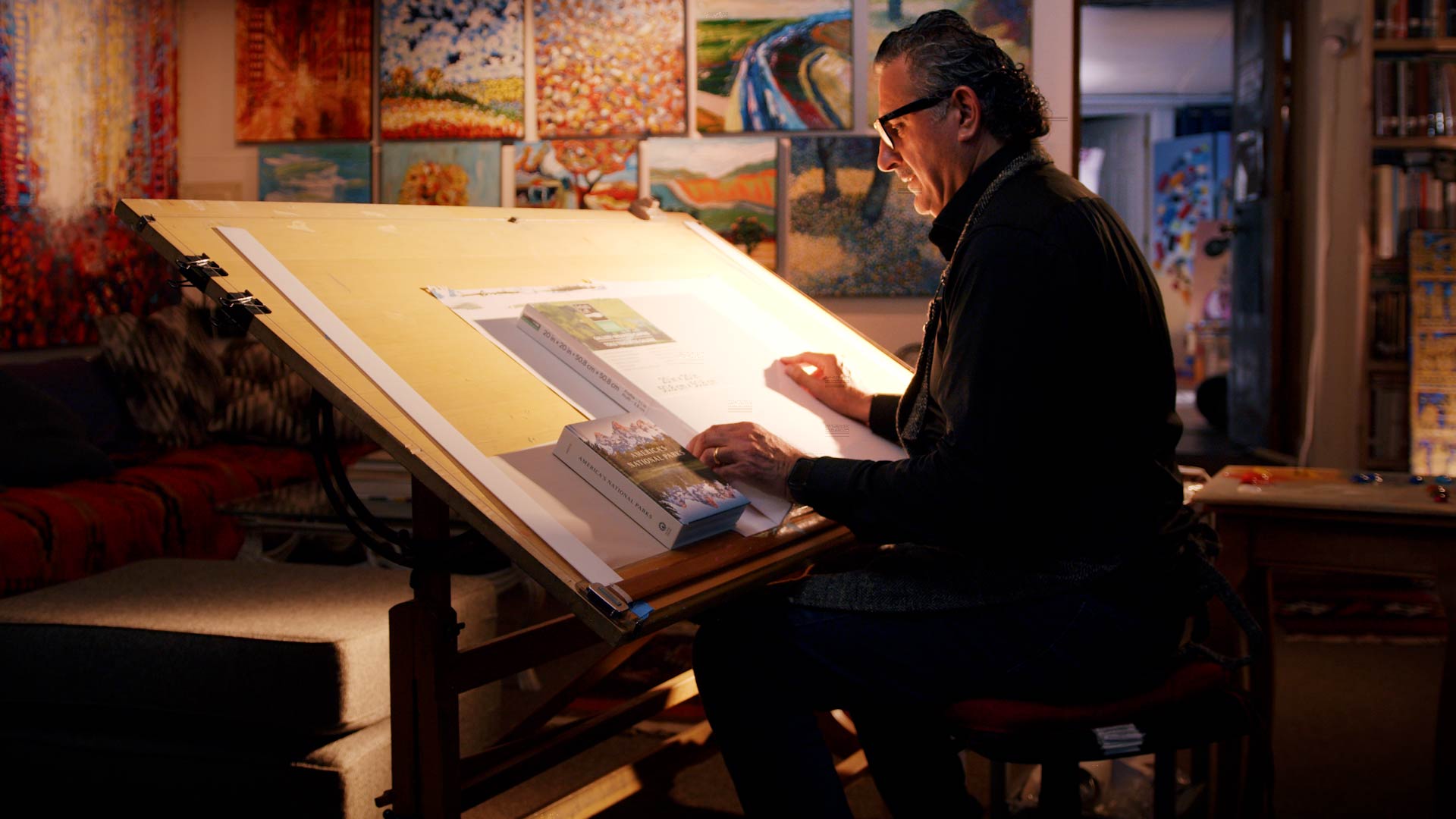 Man wearing glasses working at a tilted drafting table with books in a warmly lit art studio filled with colorful paintings.