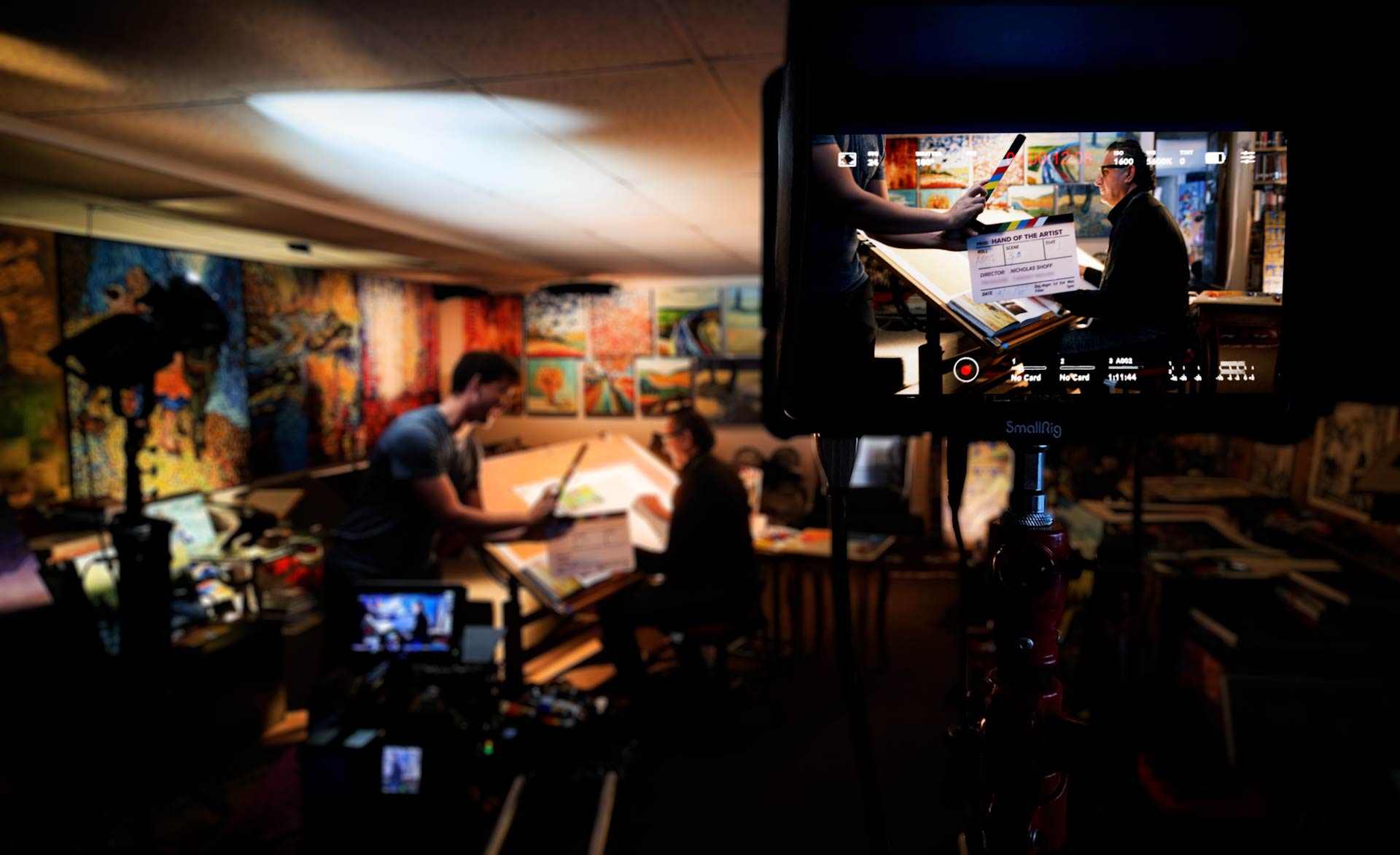 A man working on a drawing table in an art studio, filmed by a documentary crew with the monitor showing a clapperboard.