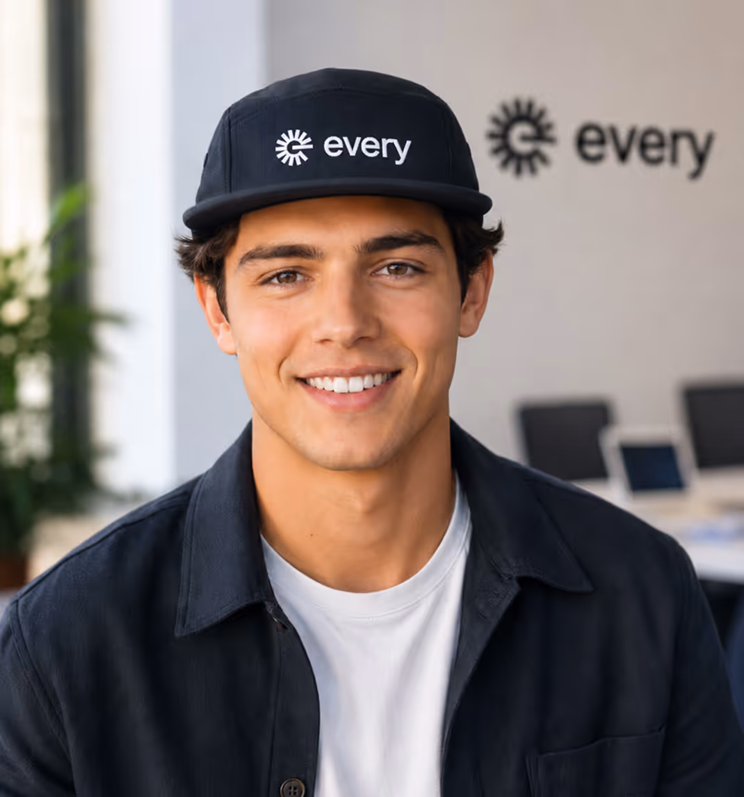 Smiling young man wearing a black cap and shirt with the word 'every' in an office setting.