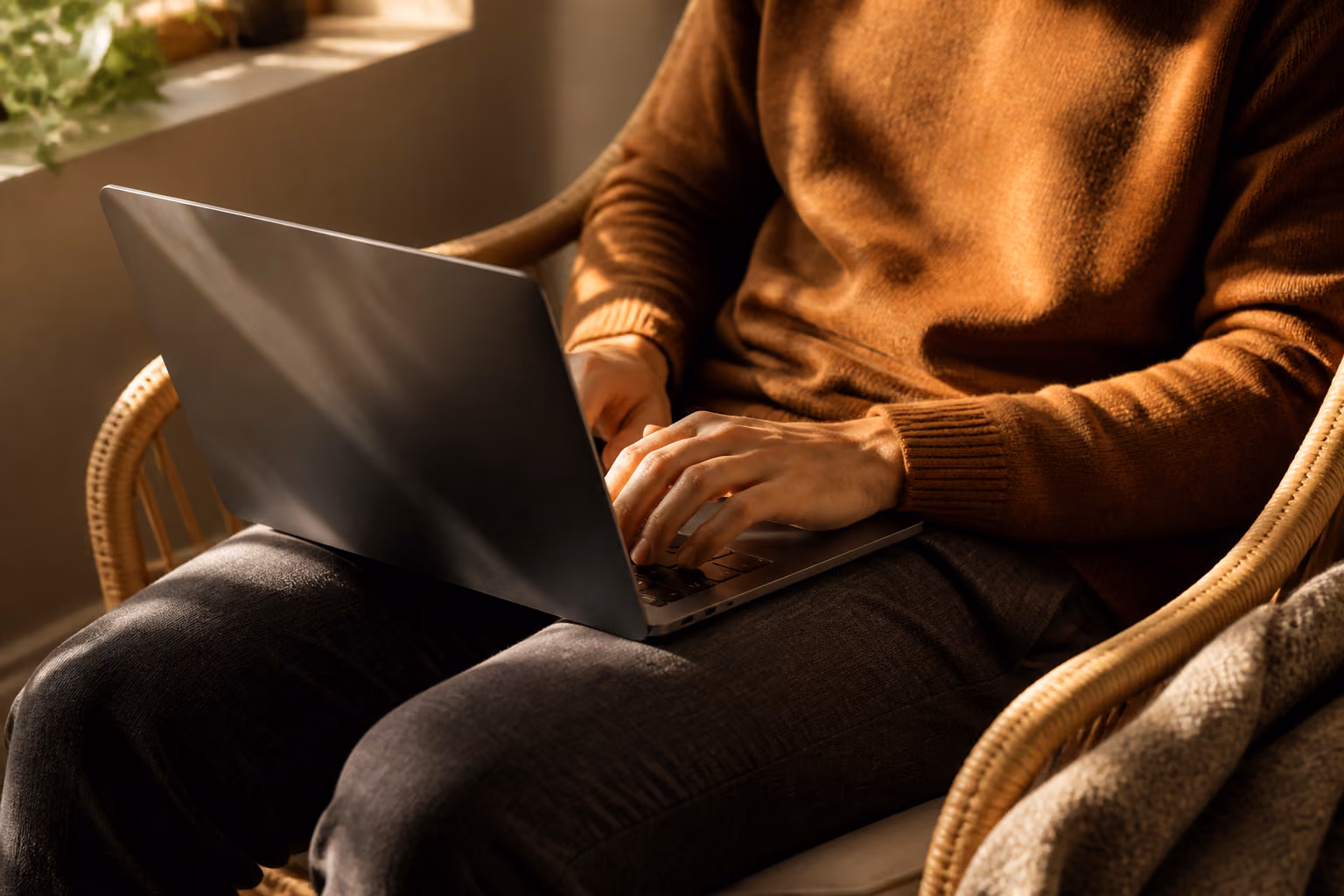 Person typing on a laptop while sitting in a wicker chair near a window with sunlight.
