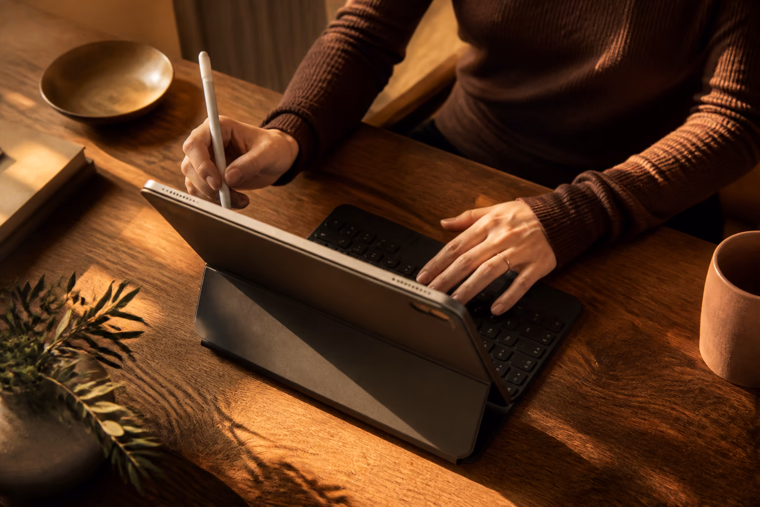 Person using a tablet with stylus and a keyboard on a wooden desk with cup and plant.