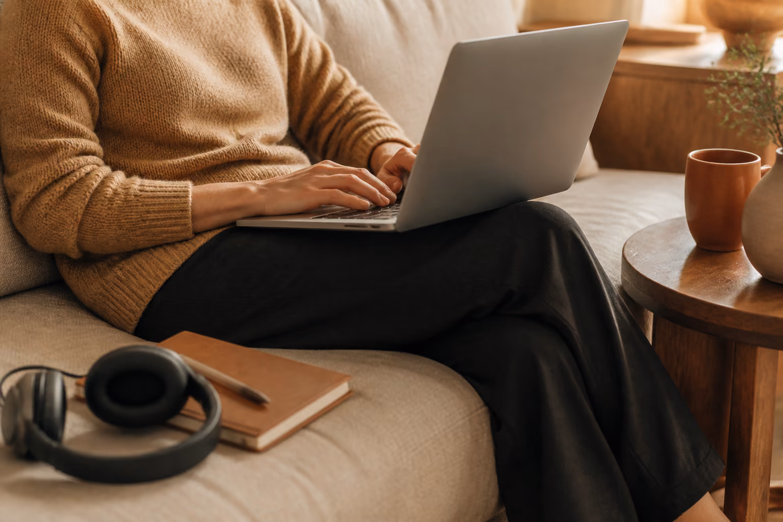 Person typing on laptop while sitting on a beige couch with headphones and notebook nearby.