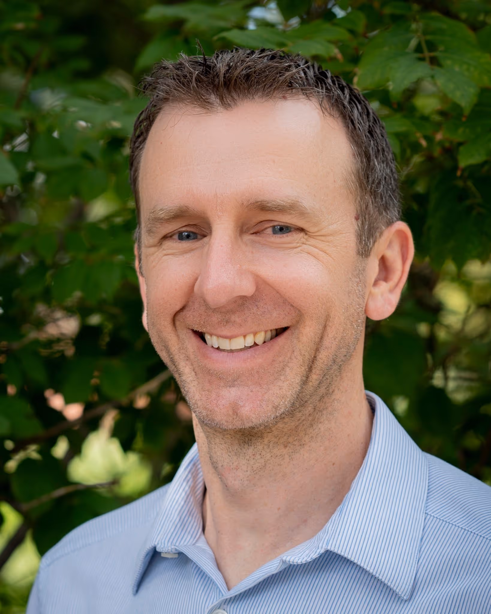 Smiling middle-aged man with short brown hair wearing a light blue striped shirt, standing outdoors with green leaves in the background.
