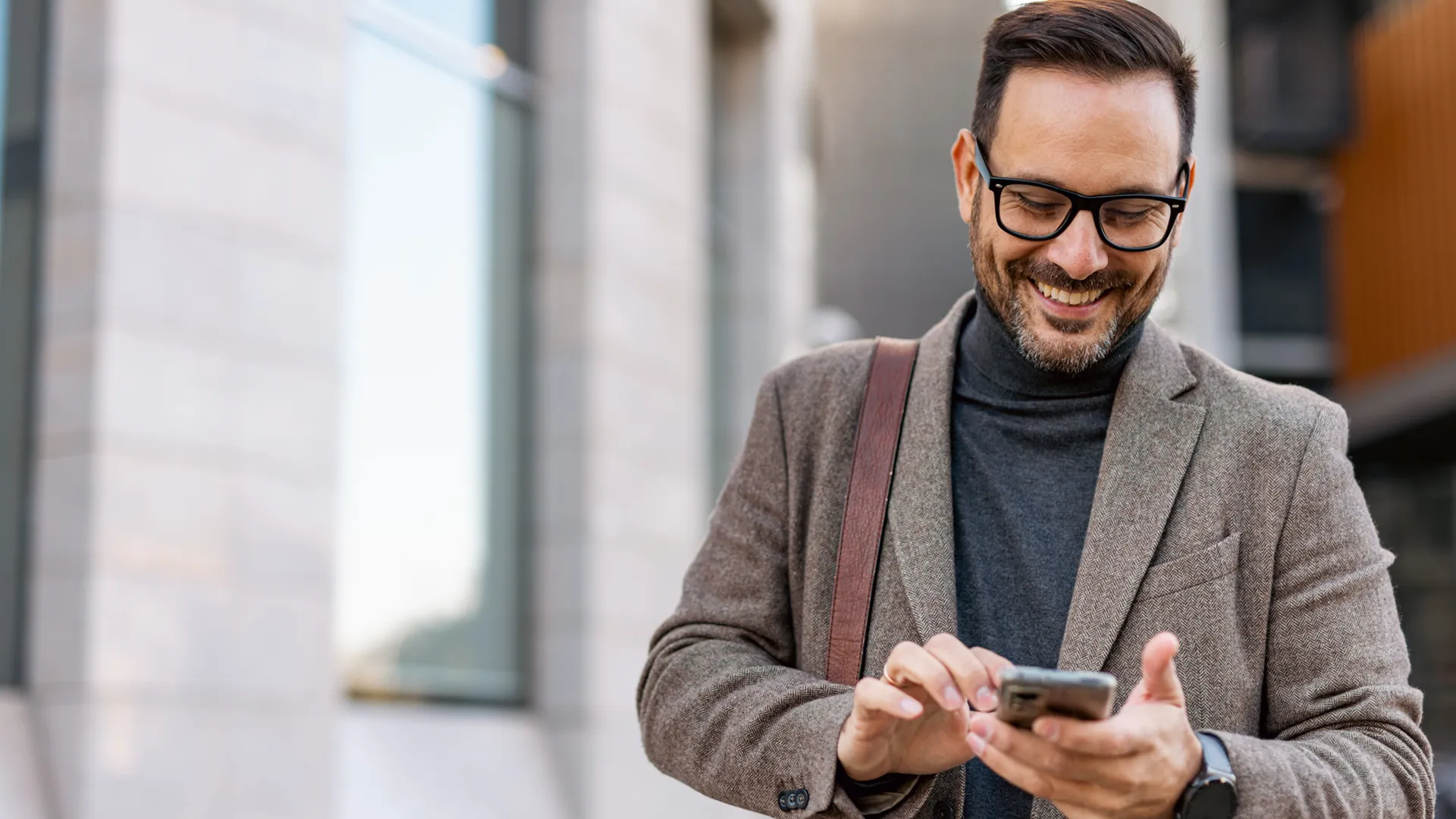 Smiling man wearing glasses and a brown jacket uses a smartphone outdoors.