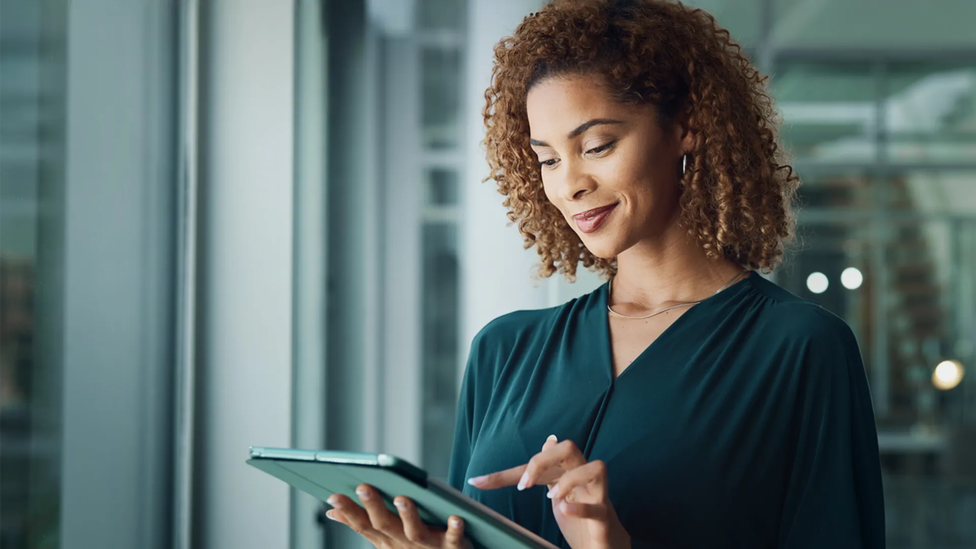 Woman with curly hair using a tablet while standing indoors near a window.
