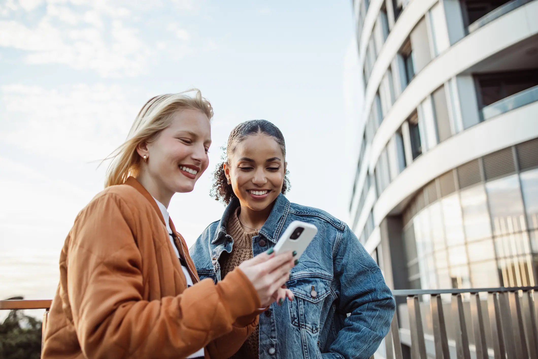 Two women smiling and looking at a smartphone outdoors near a modern curved building.