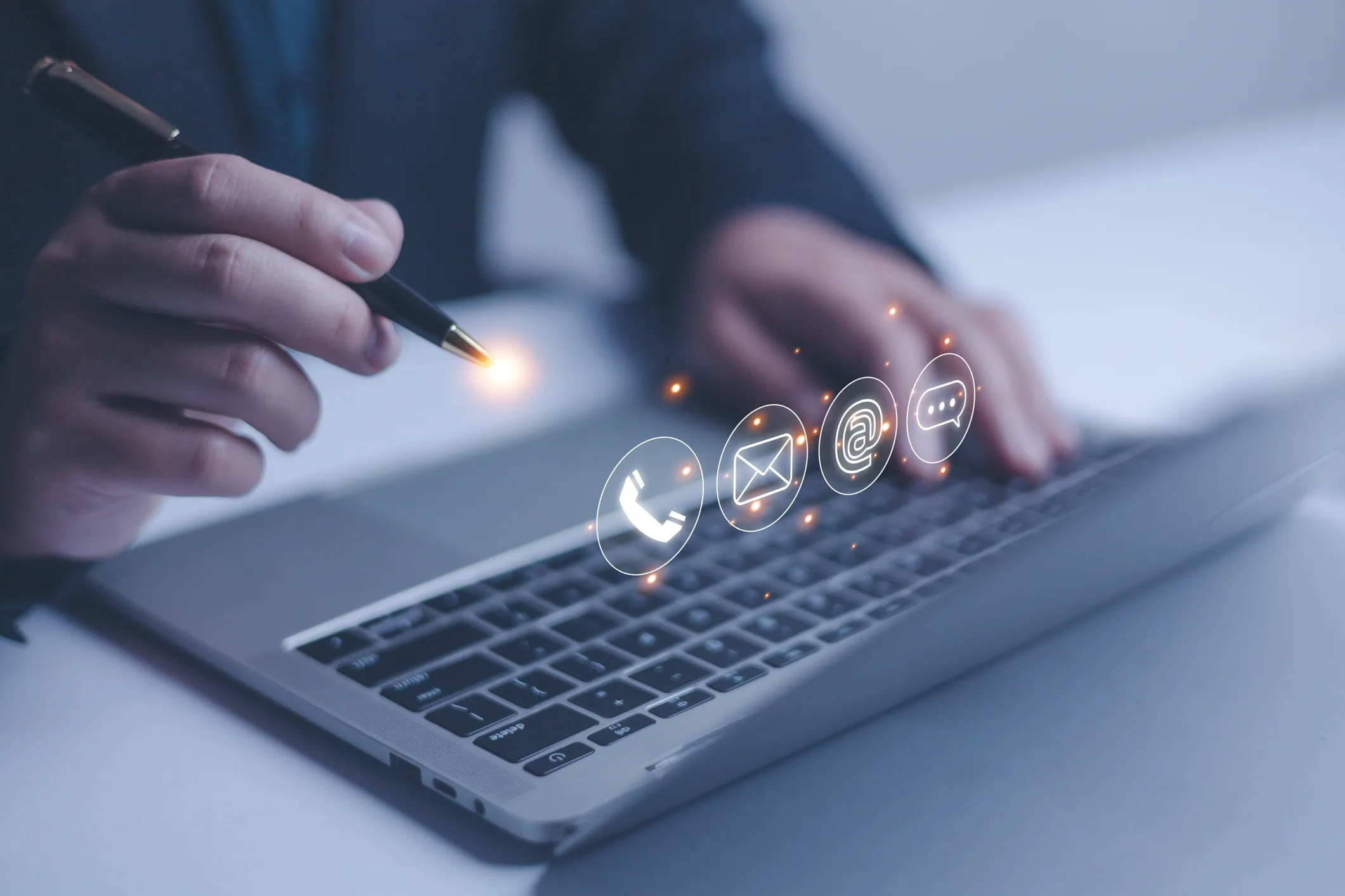 Person using a pen and laptop keyboard with glowing contact icons for phone, email, and chat hovering above.