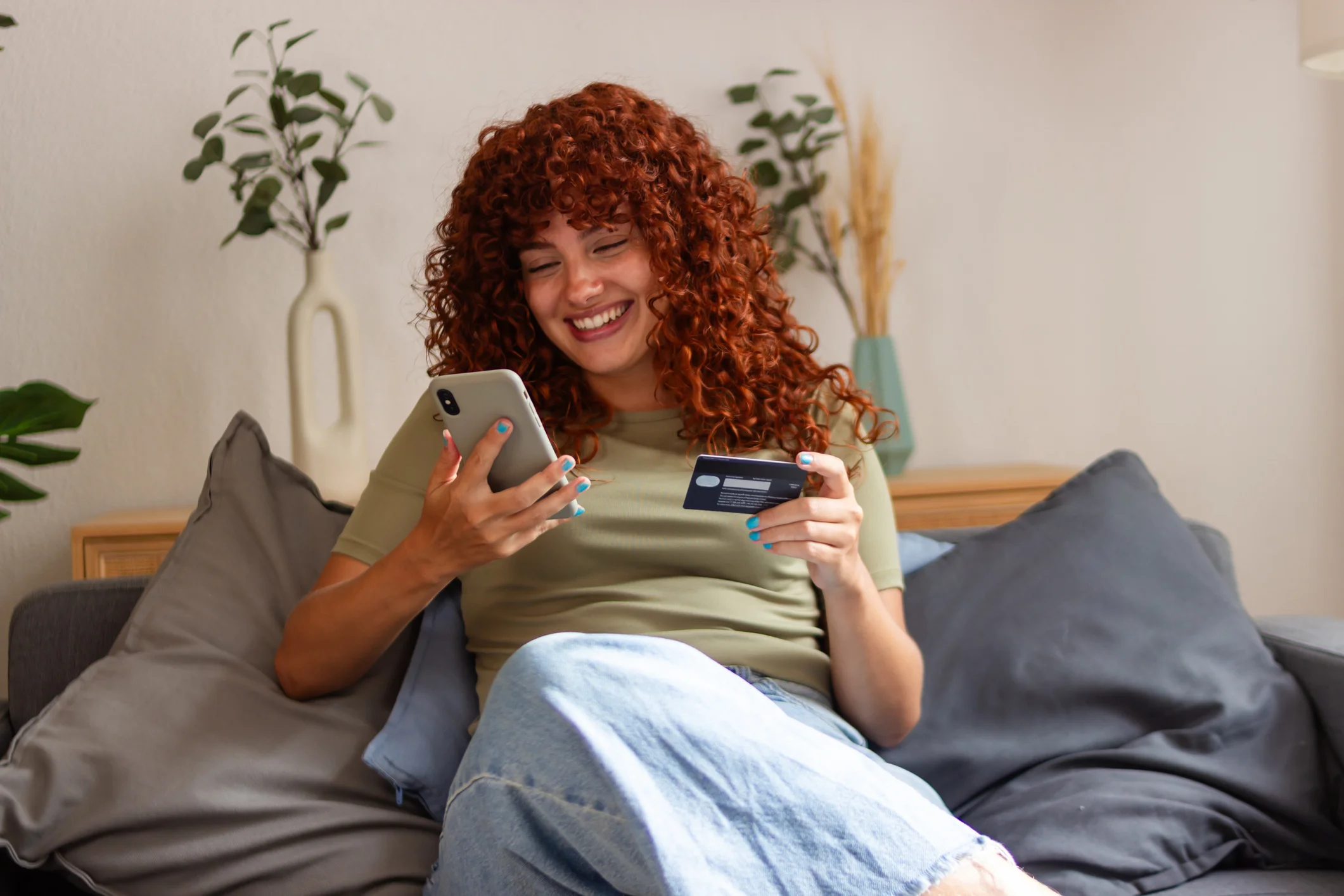 Smiling woman with curly red hair sitting on a couch using a smartphone and holding a credit card.