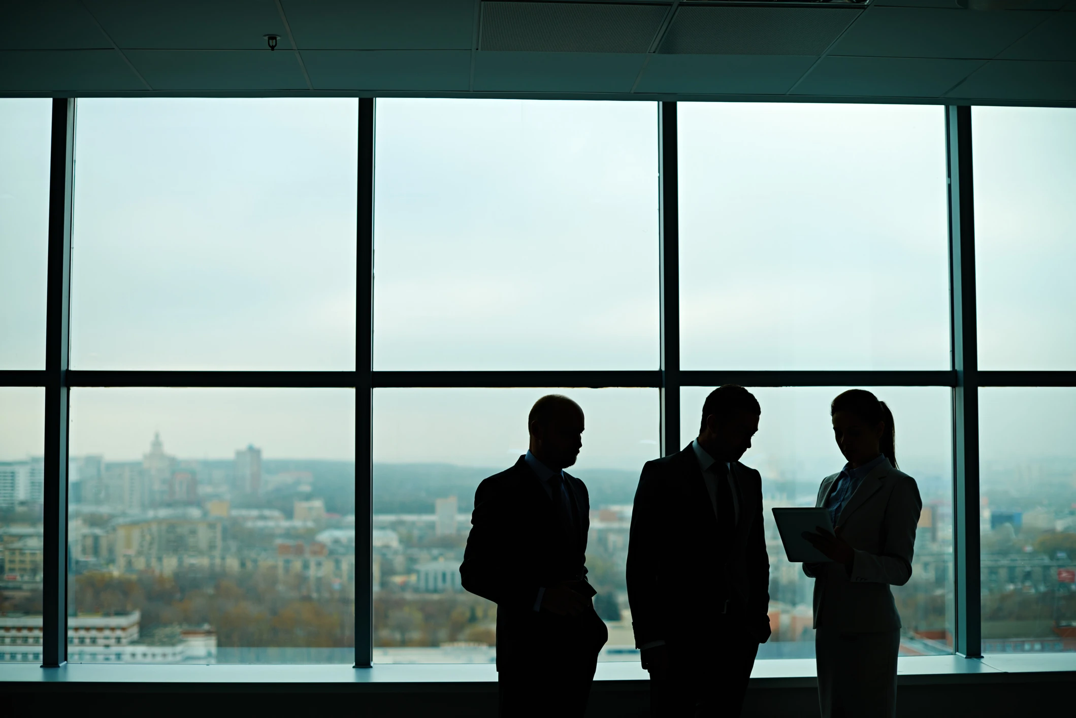 Silhouettes of three businesspeople in suits standing and discussing in front of large office windows overlooking a cityscape on a cloudy day.