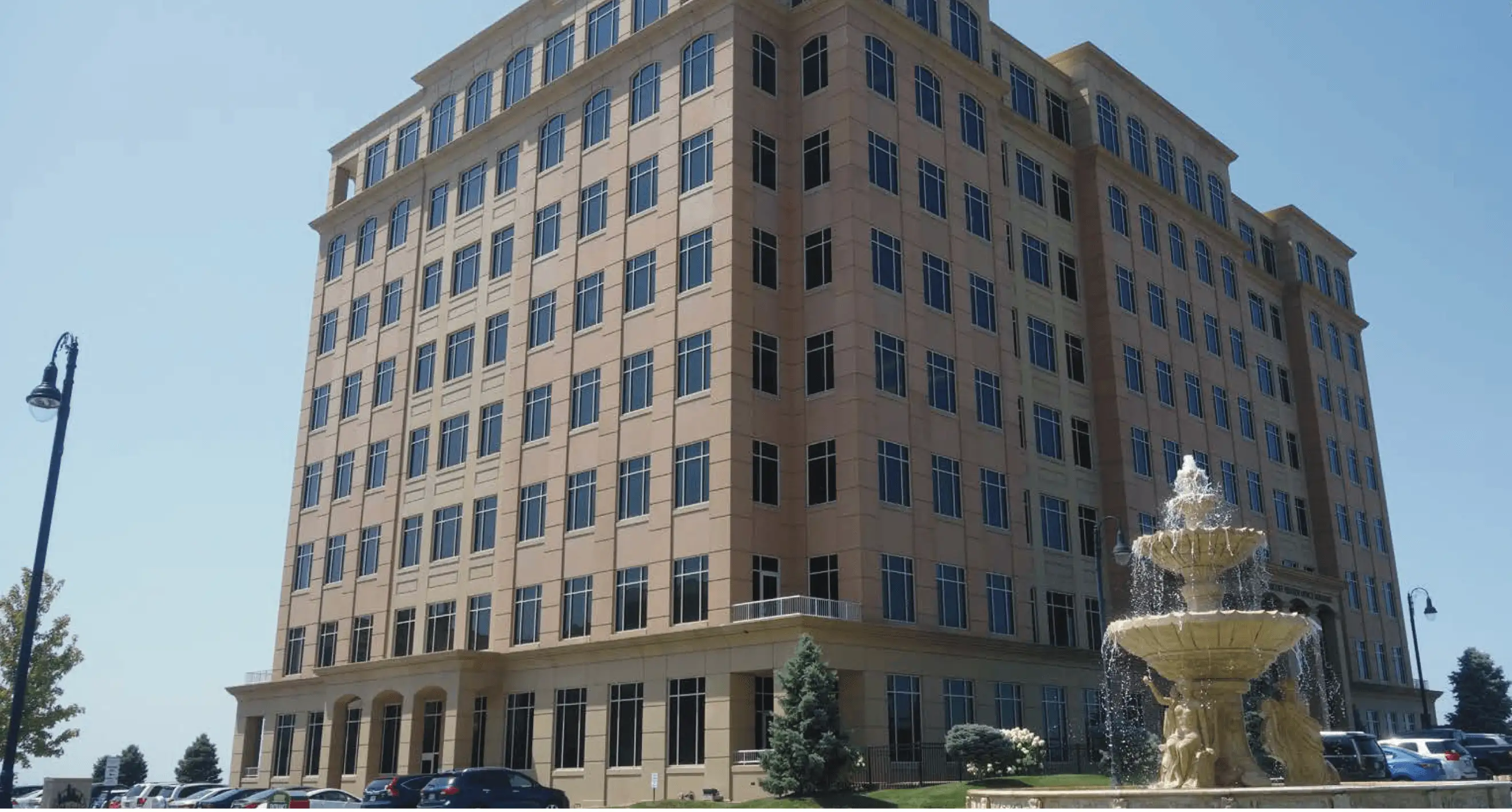 Large beige office building with many windows next to a decorative water fountain in a parking lot.