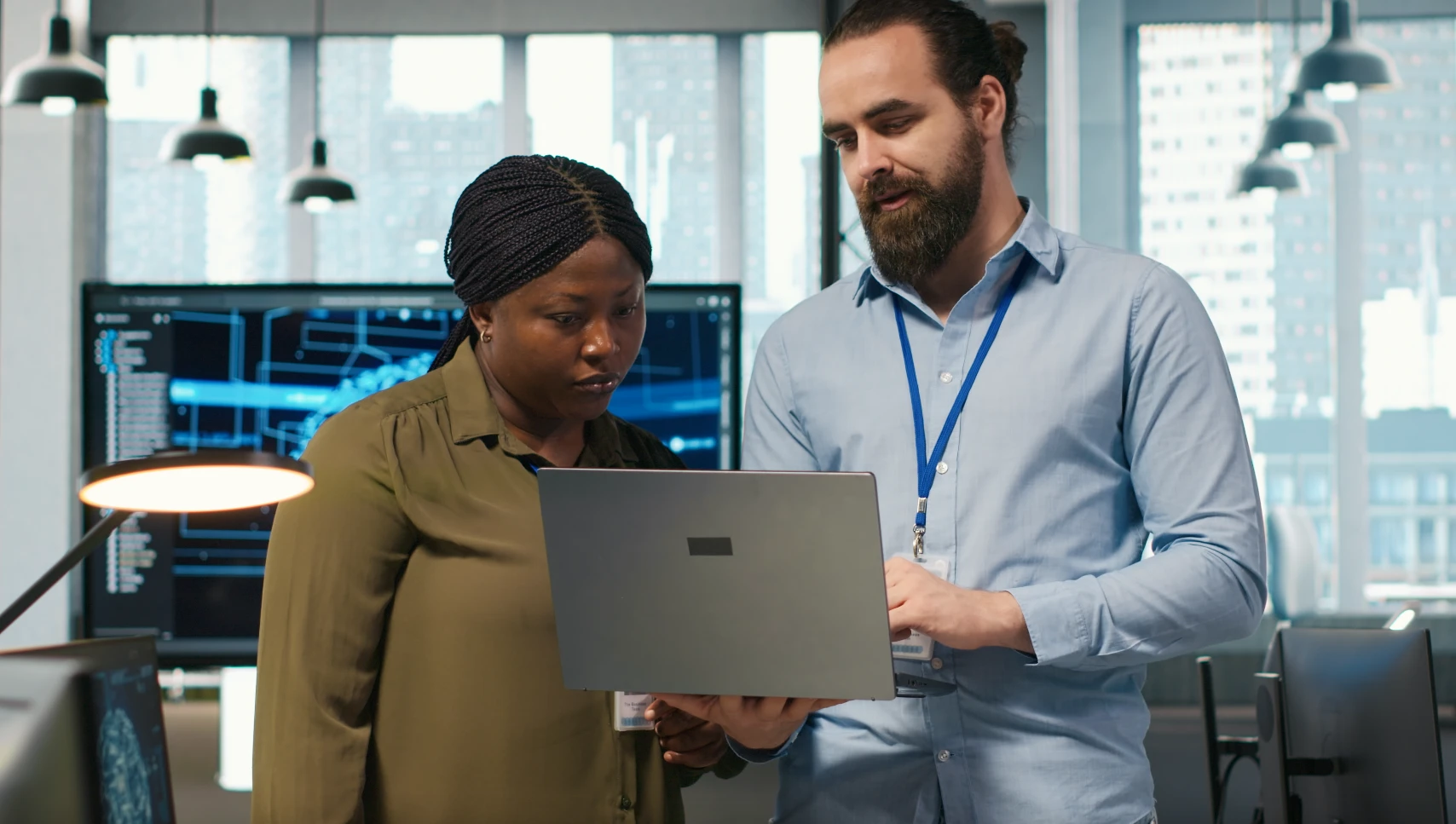Two professionals reviewing a laptop screen in a modern office with computer monitors displaying technical data in the background.