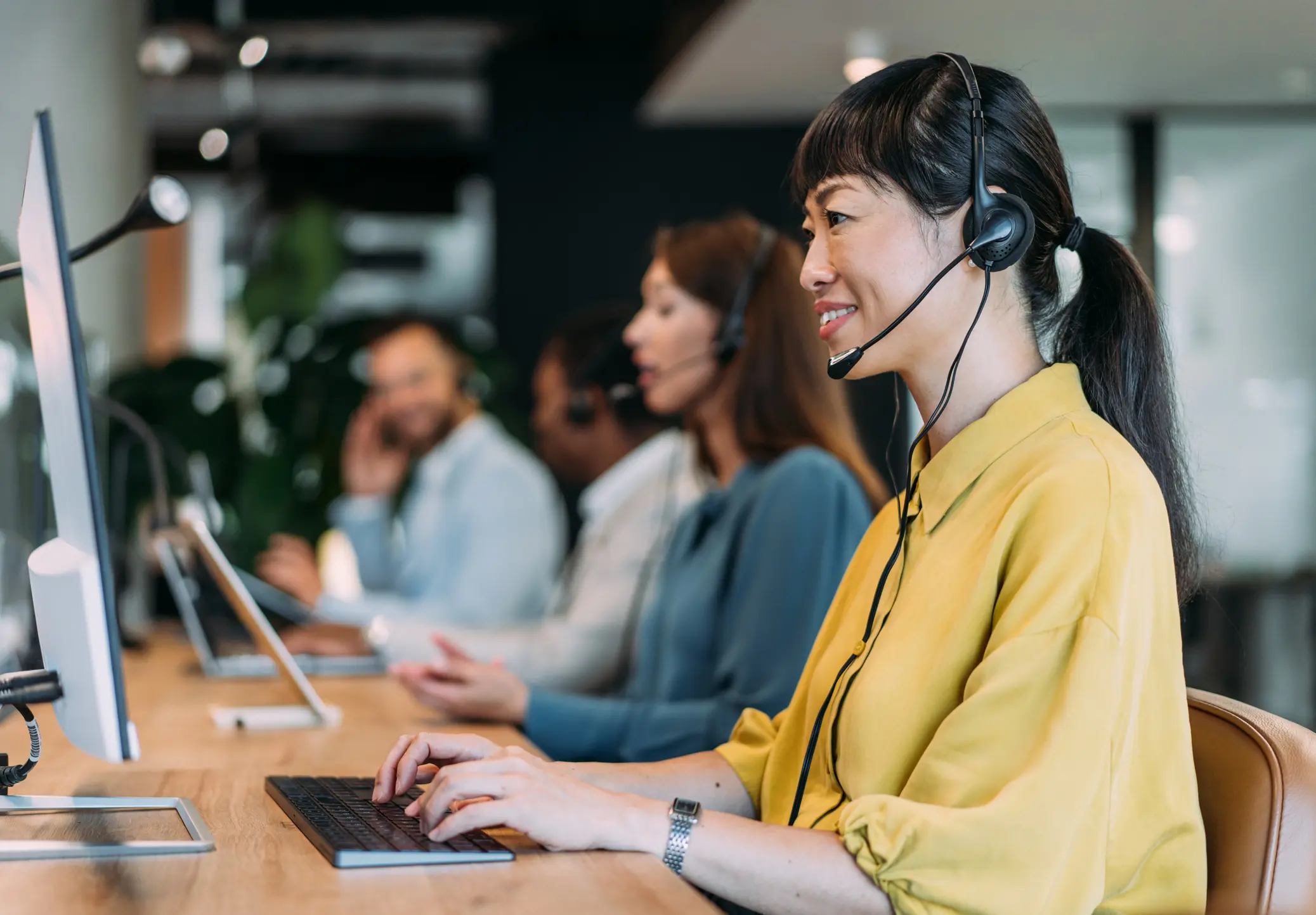 Smiling woman in a yellow shirt wearing a headset and typing on a keyboard in a call center with colleagues in the background.