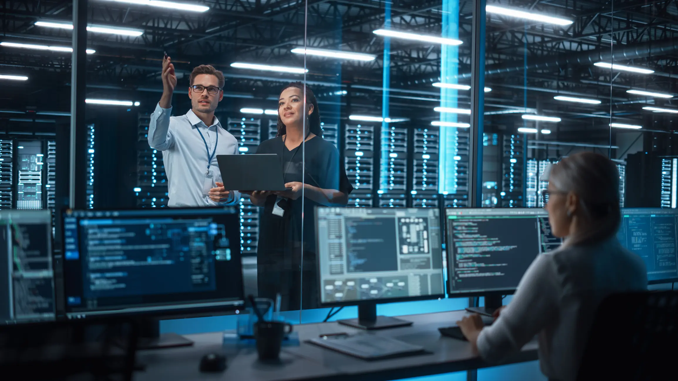 Two IT professionals standing and discussing data on a laptop with a woman working on multiple monitors displaying code in a server room.
