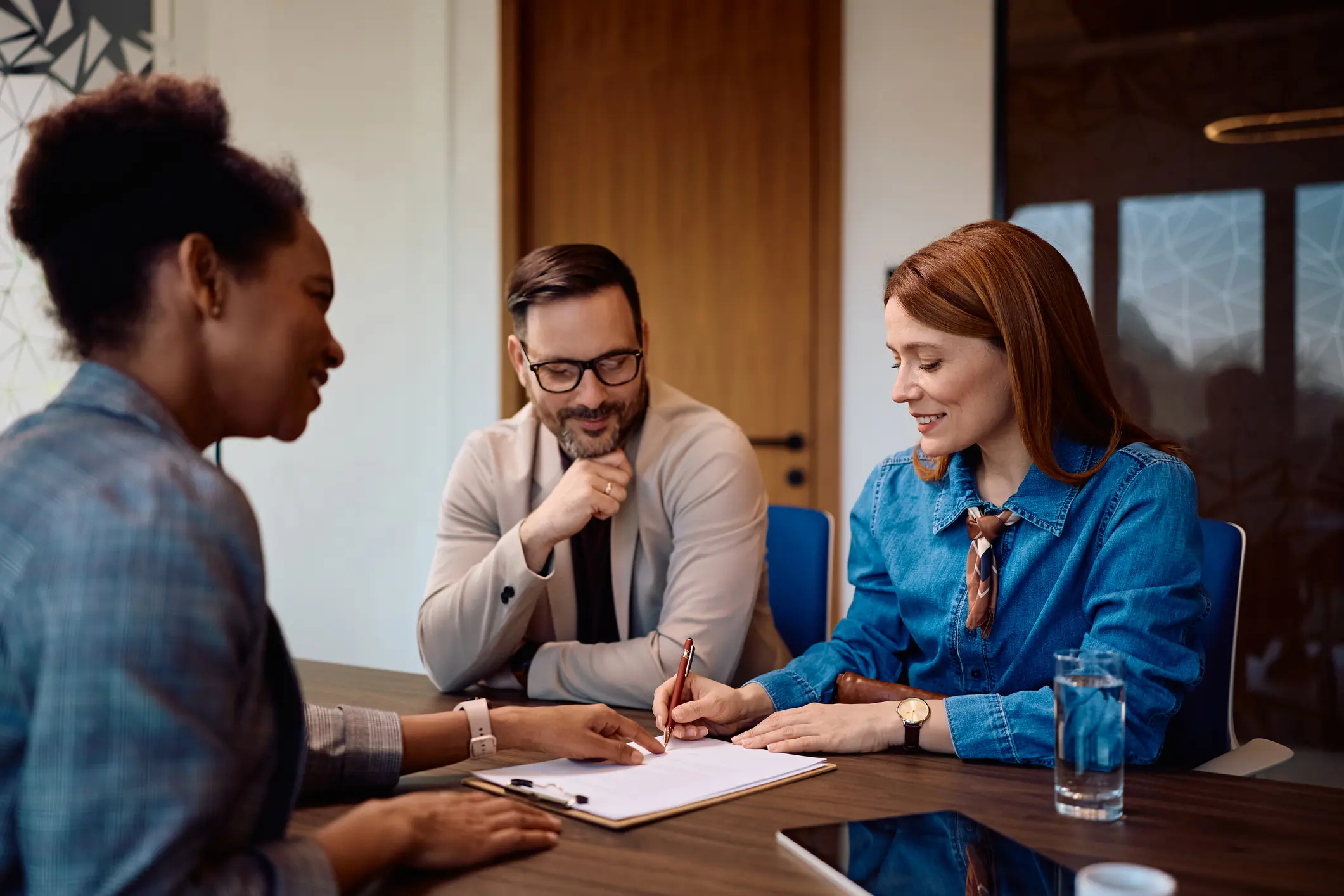 Three colleagues sitting at a table, one woman is signing a document while the others watch and smile.