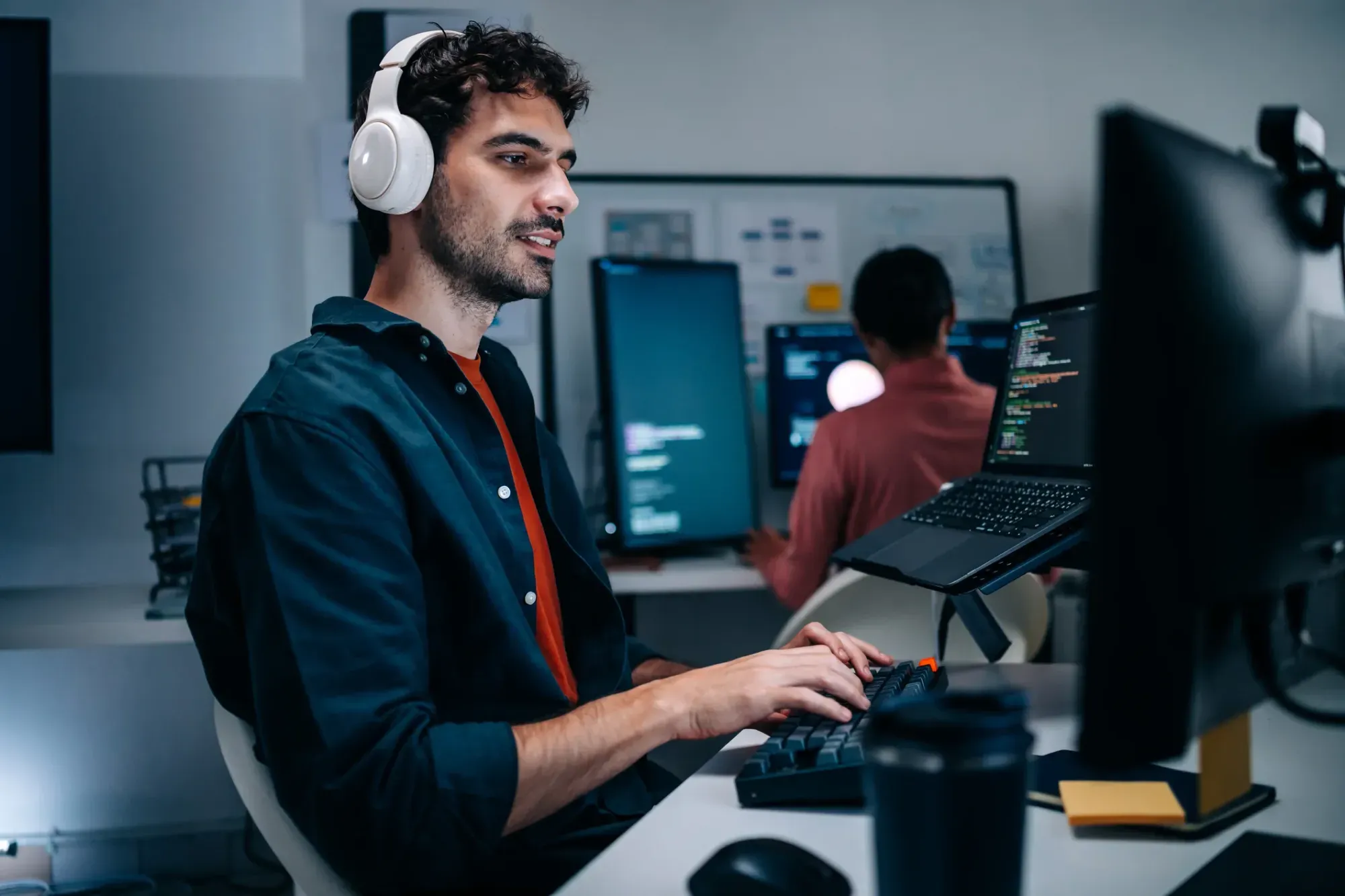 Man wearing white headphones typing on a keyboard in front of multiple computer screens displaying code in a modern office setting.