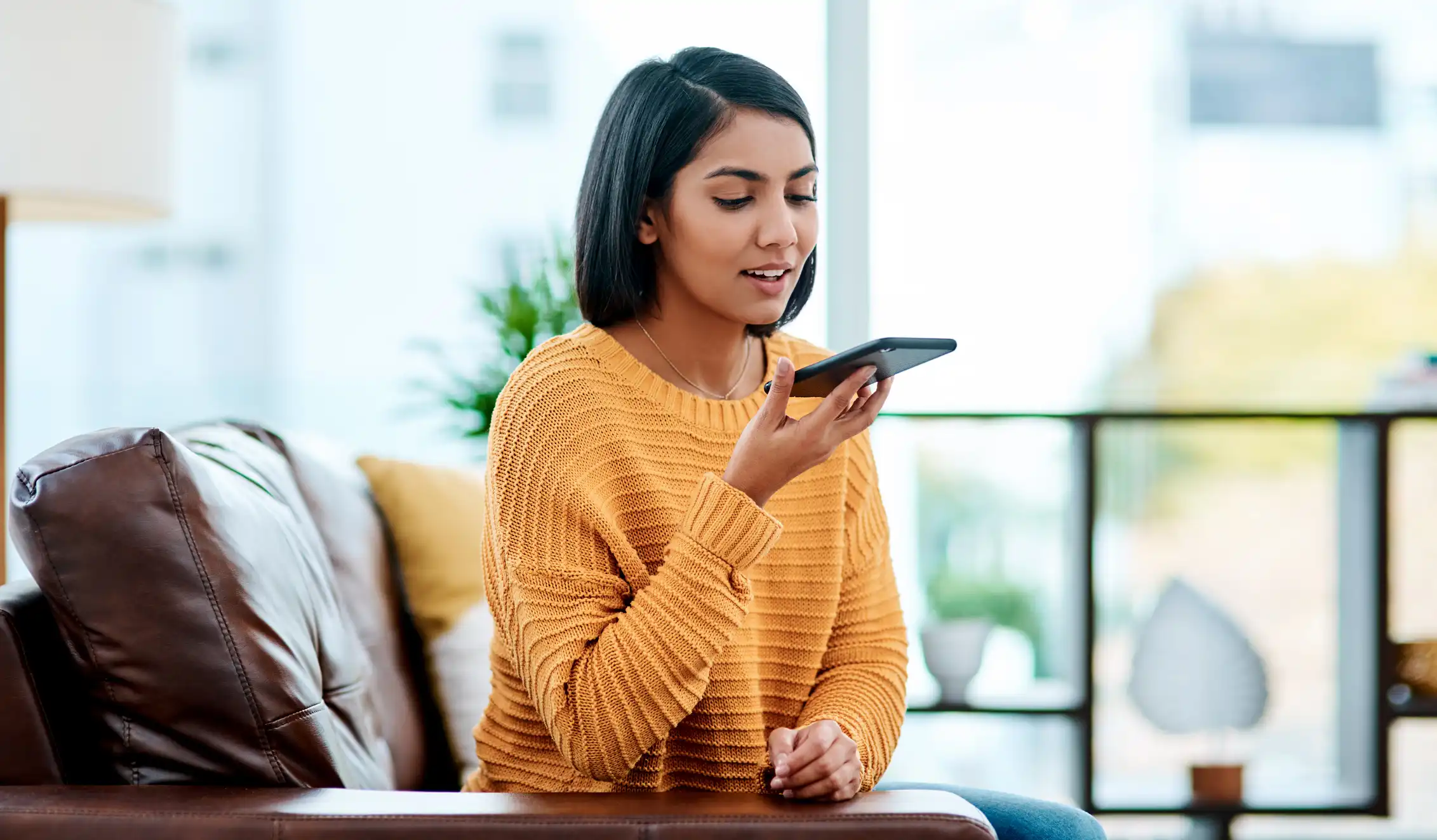 Woman using her smartphone's voice assistant while sitting on a sofa in a bright living room.