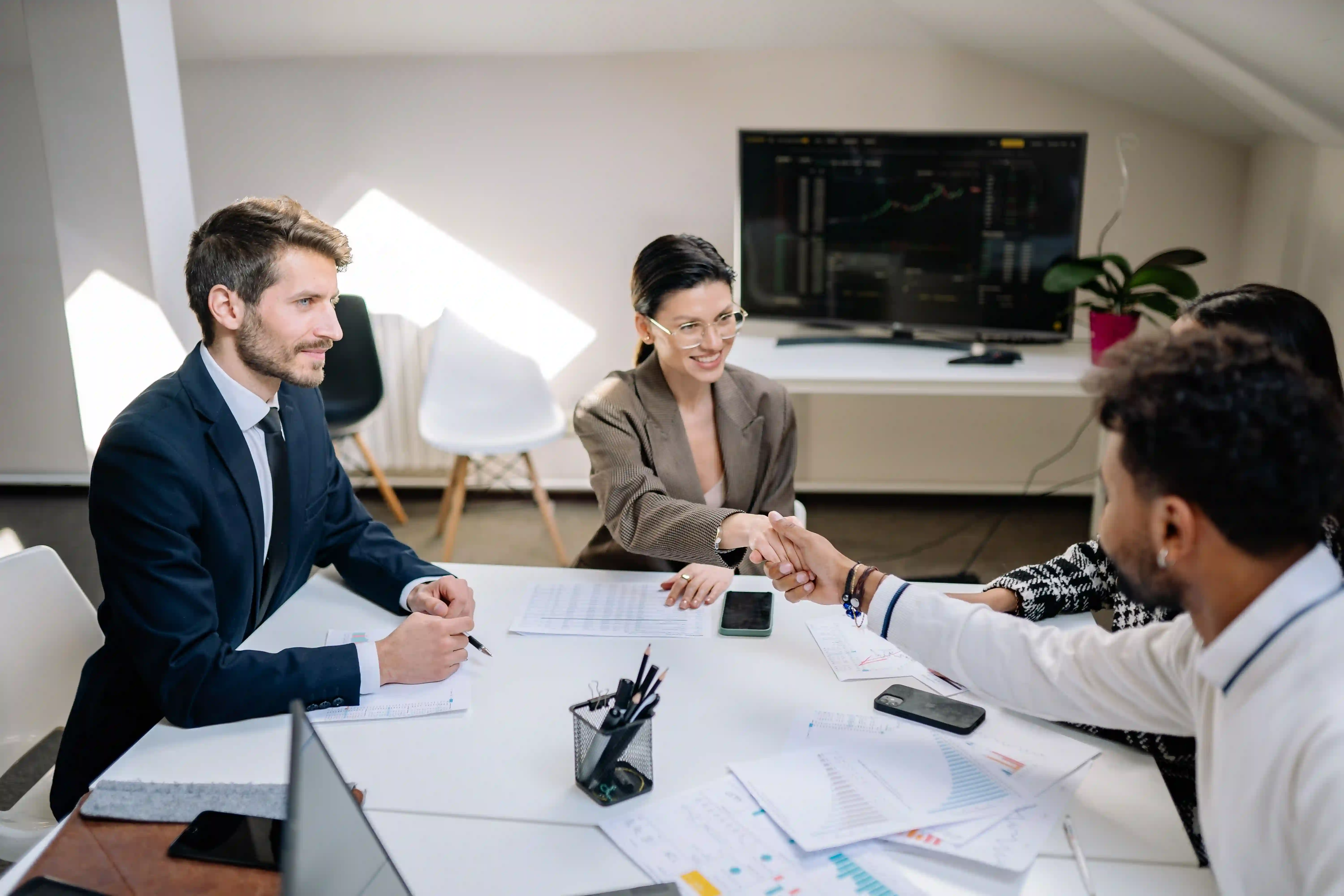 Three business professionals meeting around a table, reviewing documents and shaking hands in an office.