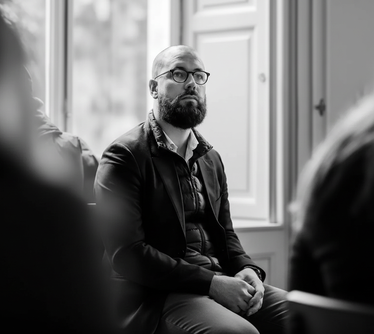Un homme barbu portant des lunettes et une veste sombre assis en posture attentive dans une pièce avec une fenêtre à l'arrière-plan.
