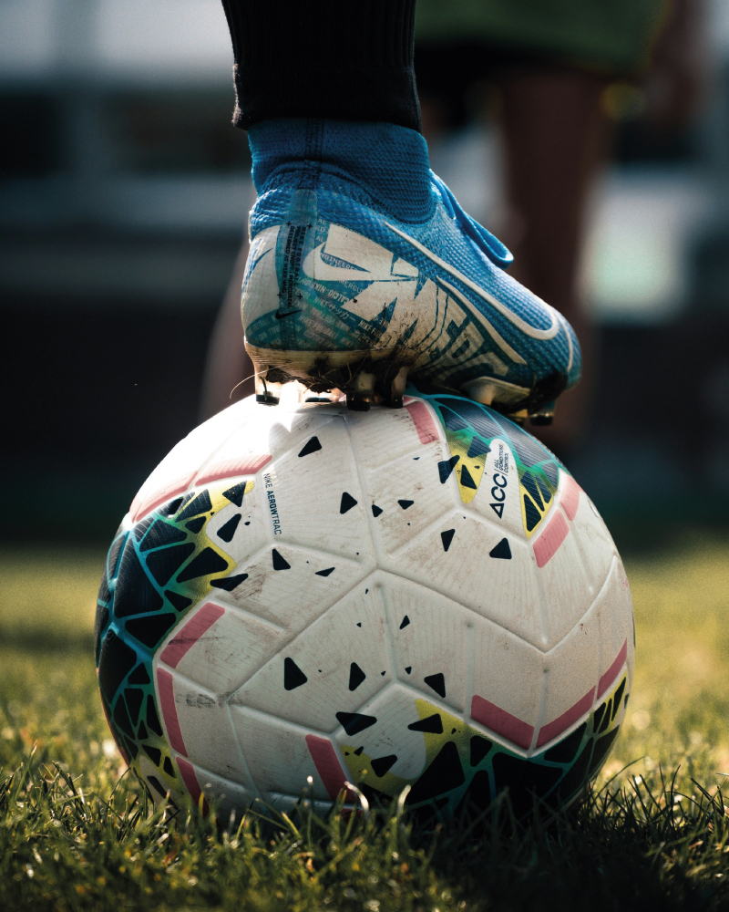 Close-up of a blue cleat stepping on a white and colorful soccer ball on grass.
