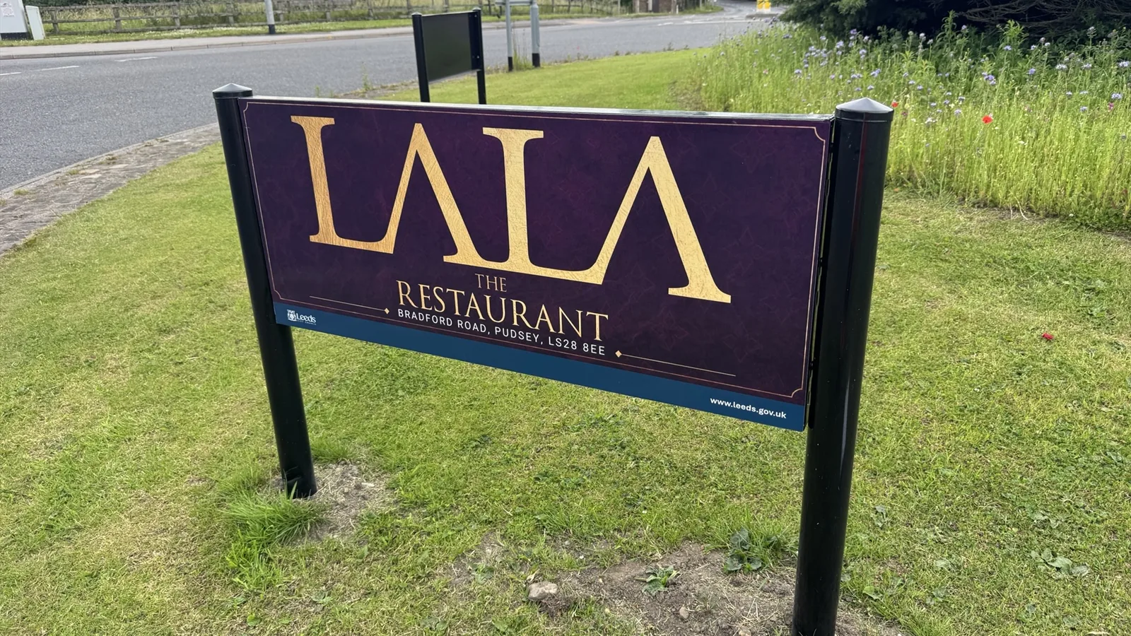 Lala The Restaurant roundabout sponsorship sign on a grass verge beside Bradford Road in Pudsey, Leeds, showing the restaurant's gold logo and address on a deep purple panel.
