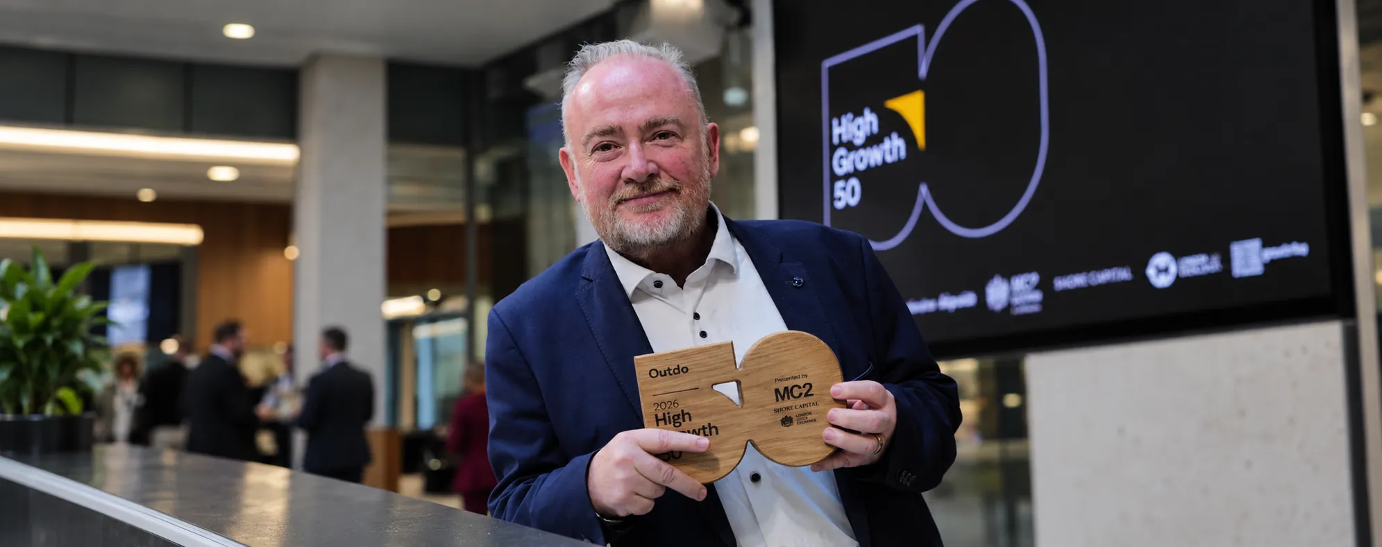 Outdo CEO Mike Brennan holding the wooden 2026 High Growth 50 award at the London Stock Exchange, with the High Growth 50 logo on a screen behind him.