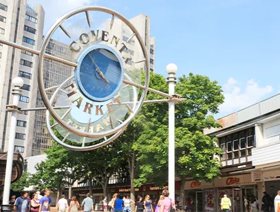 Circular Coventry Market clock sign above shoppers, with a tall building and trees behind
