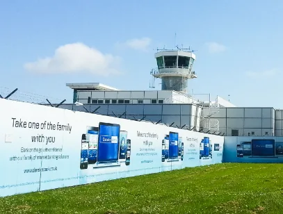 Airport control tower behind a long fence hoarding with advertising, seen across grass near Derry