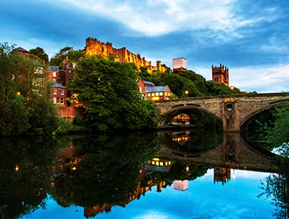 Stone bridge over the River Wear in Durham at dusk, with the castle and cathedral above the trees