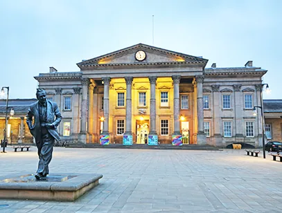 Floodlit columned railway station facade with a statue in the square at dusk in Huddersfield