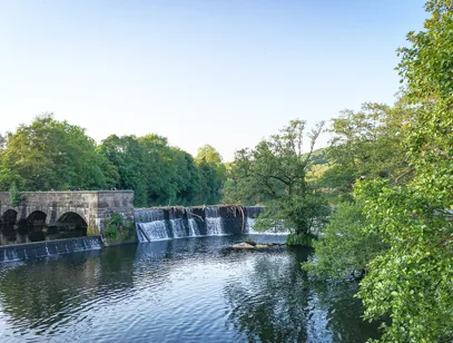 Stone weir and arched bridge on a calm river surrounded by green trees in the High Peak