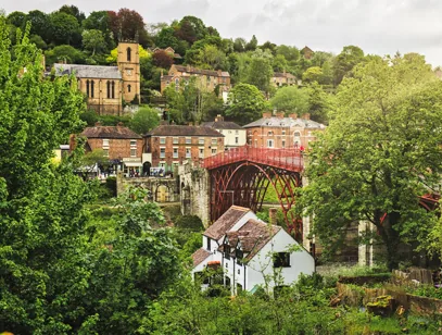 Red iron bridge crossing a wooded gorge with houses and a church on the hillside in Shropshire