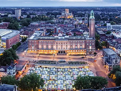 Norwich City Hall and clock tower lit at dusk above the striped market stalls, with the cathedral beyond
