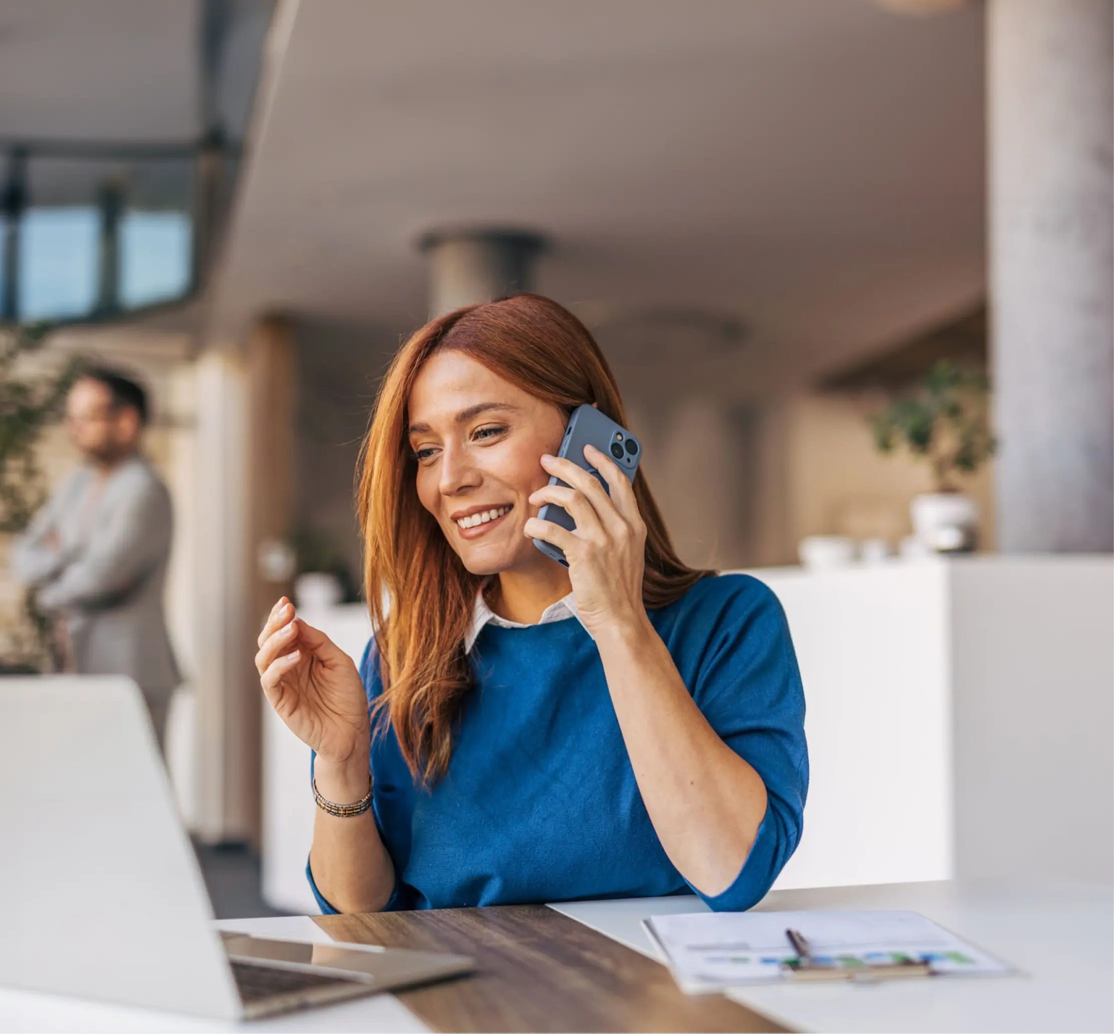 Frau telefoniert lächelnd und arbeitet an einem Laptop in einem modernen Büro.
