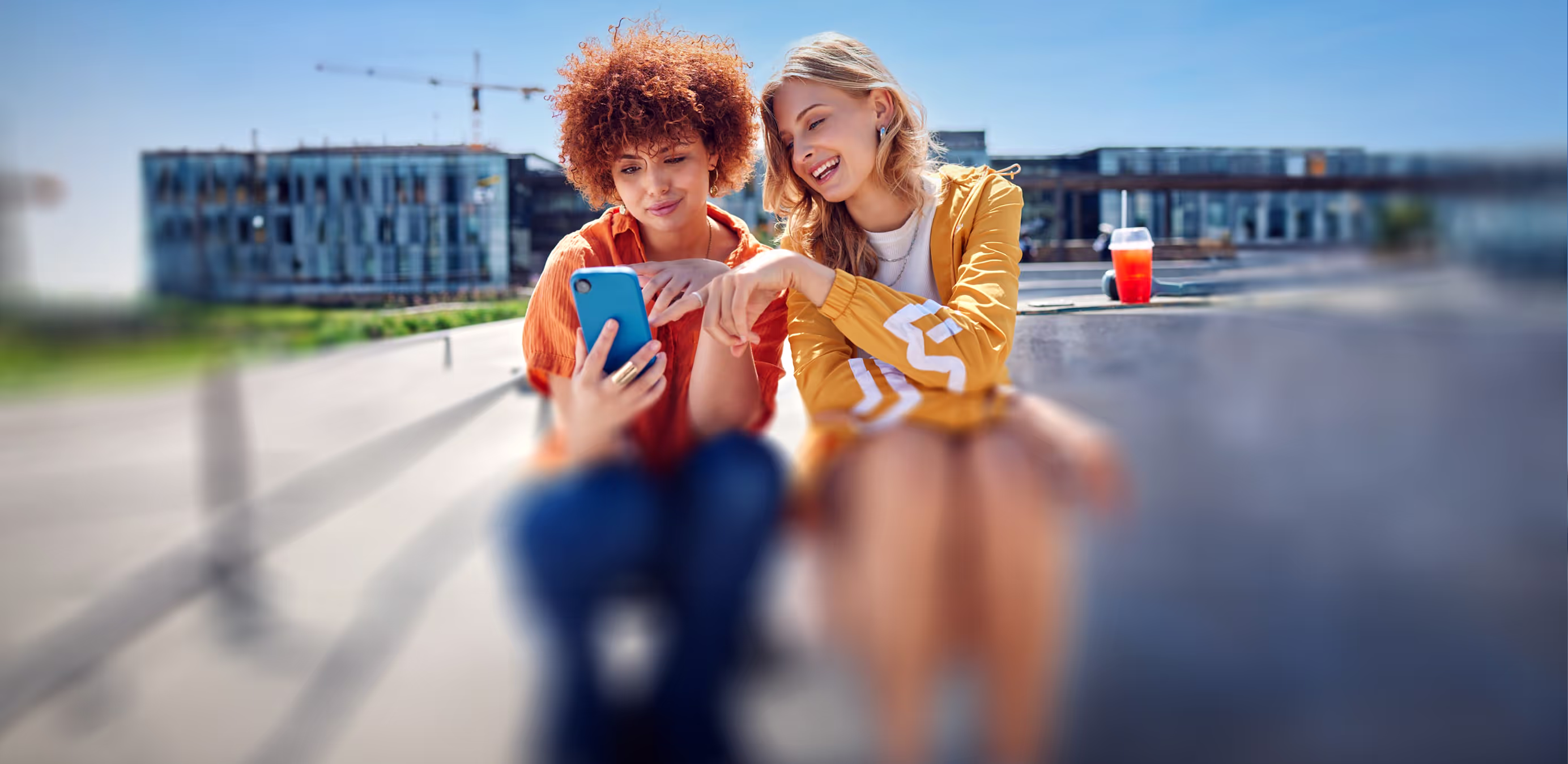 Zwei junge Frauen sitzen draußen und schauen gemeinsam auf ein Smartphone, eine trägt eine orange Bluse, die andere ein gelbes Oberteil.