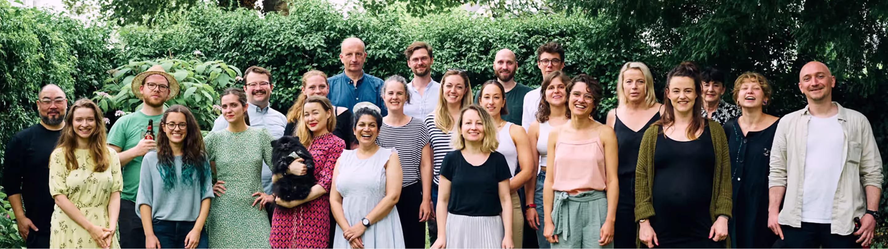 Eine Gruppe von 23 Personen, meist lächelnd, steht vor einem üppigen grünen Hintergrund, eine Frau hält einen kleinen schwarzen Hund.