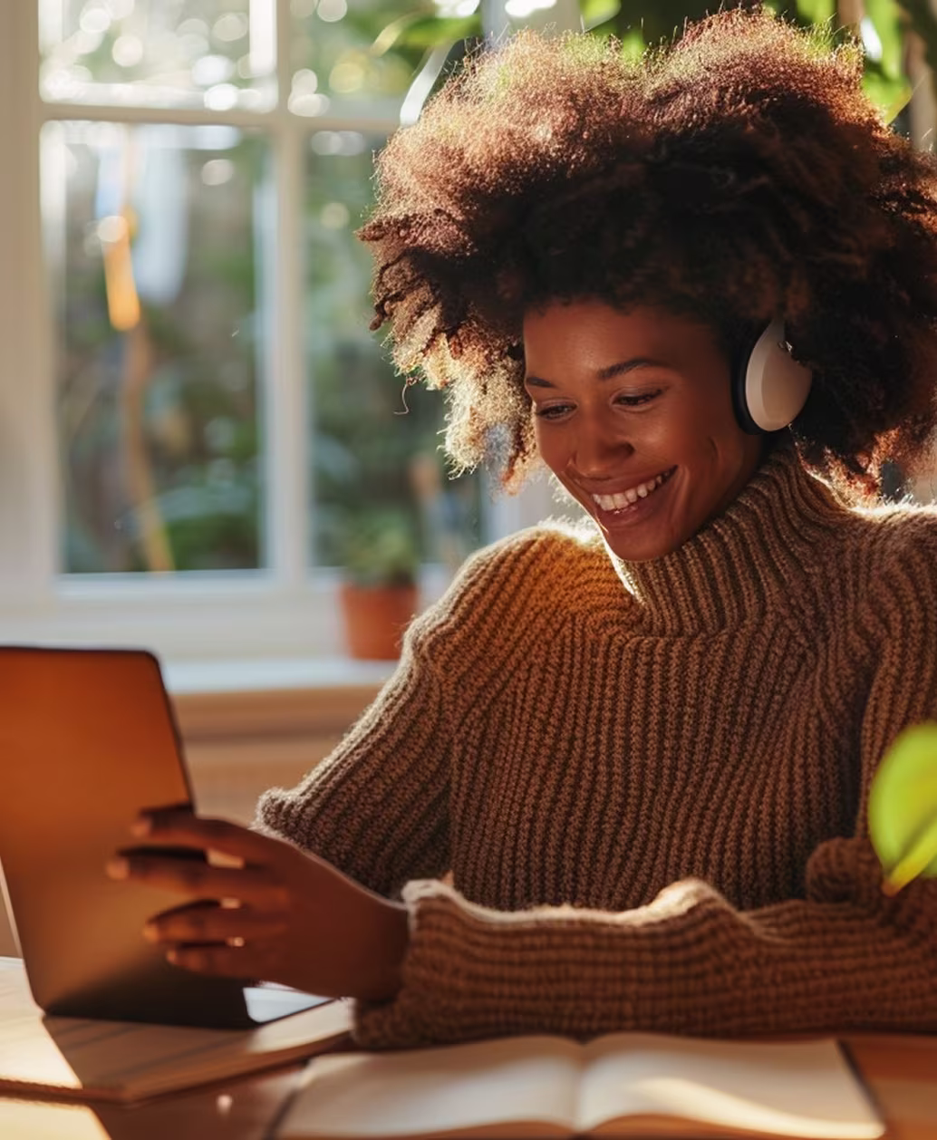 Frau mit Kopfhörern und Wollpullover lächelnd, während sie ein Tablet benutzt und an einem Tisch mit Buch sitzt.