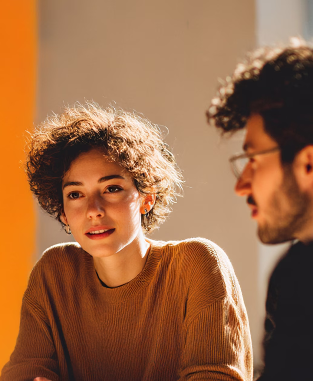 Zwei Personen unterhalten sich, eine Frau mit kurzen lockigen Haaren und braunem Pullover ist im Fokus.