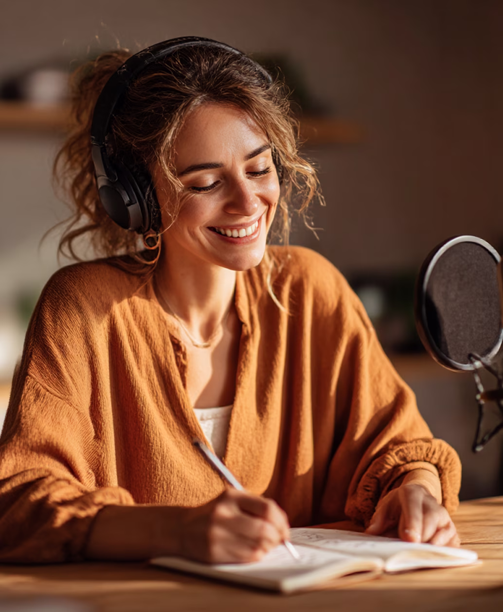Frau mit Kopfhörern lächelt und schreibt in ein Notizbuch an einem Tisch mit Mikrofon.
