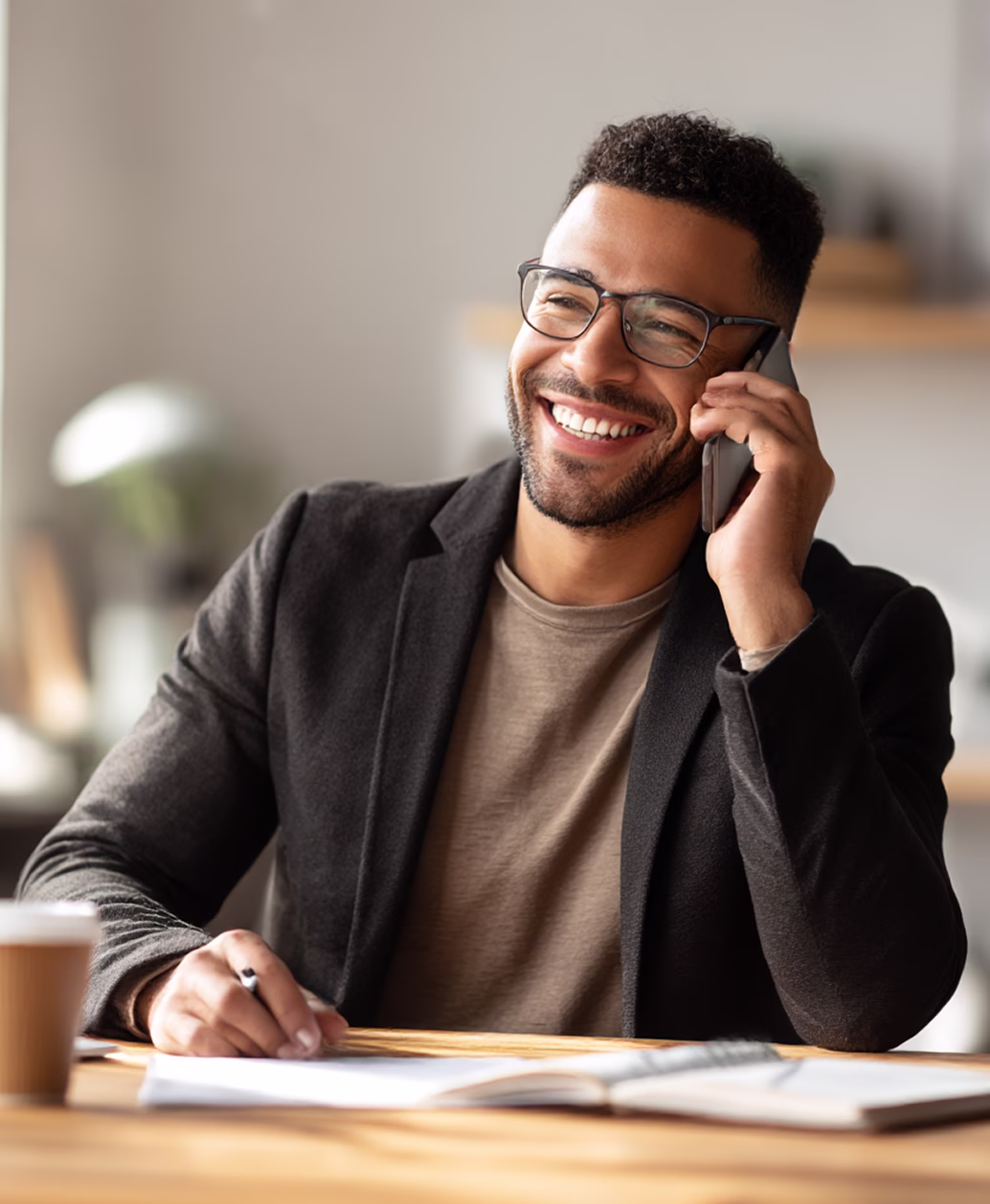 Lächelnder Mann mit Brille spricht am Telefon und macht sich Notizen an einem Holztisch.