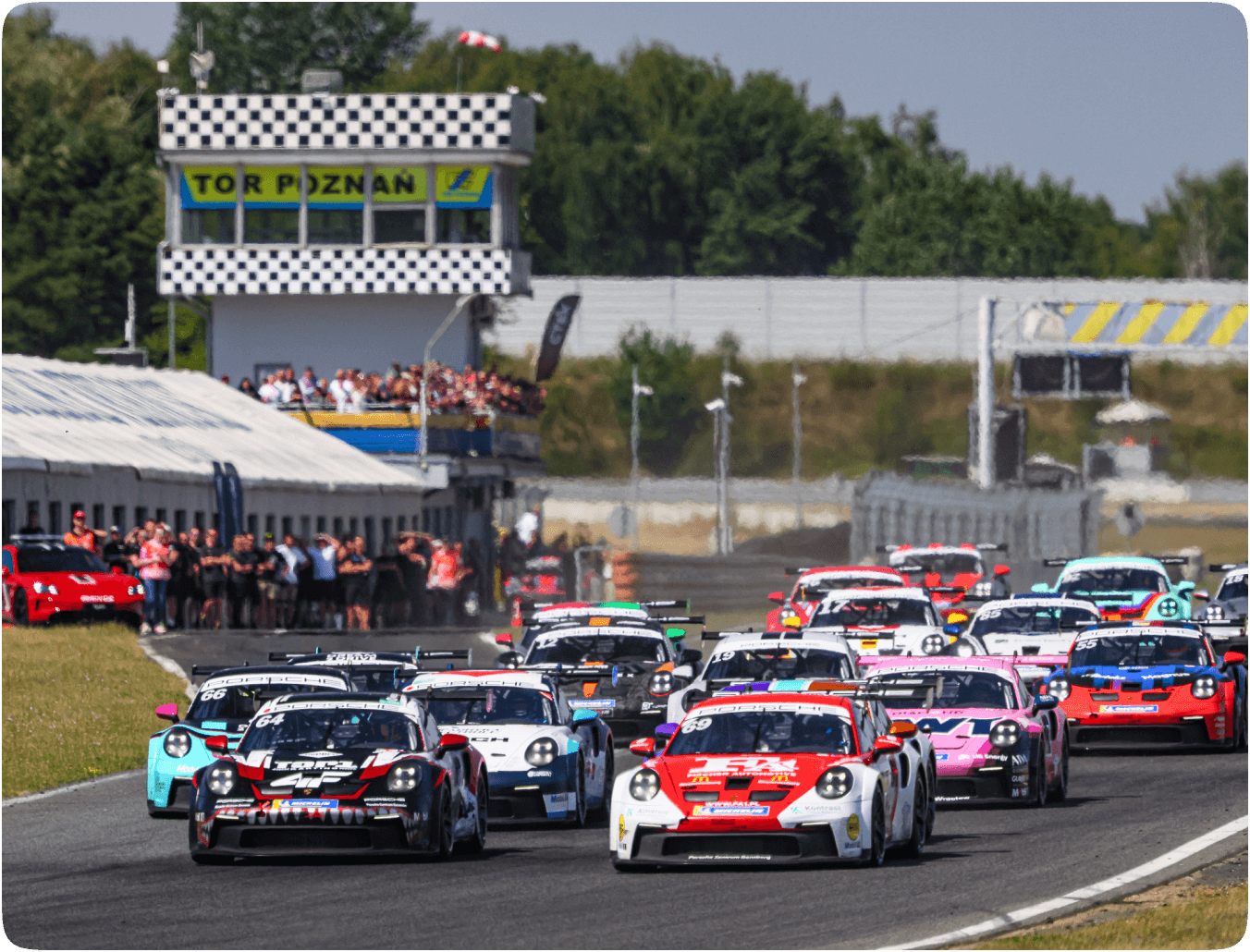 **Alt Text:** Start wyścigu Porsche Sprint Challenge Central Europe na Torze Poznań – czołówka stawki z Fabianem Dybionką na prowadzeniu, tłumy kibiców na trybunach, słoneczna pogoda i intensywna rywalizacja na starcie.