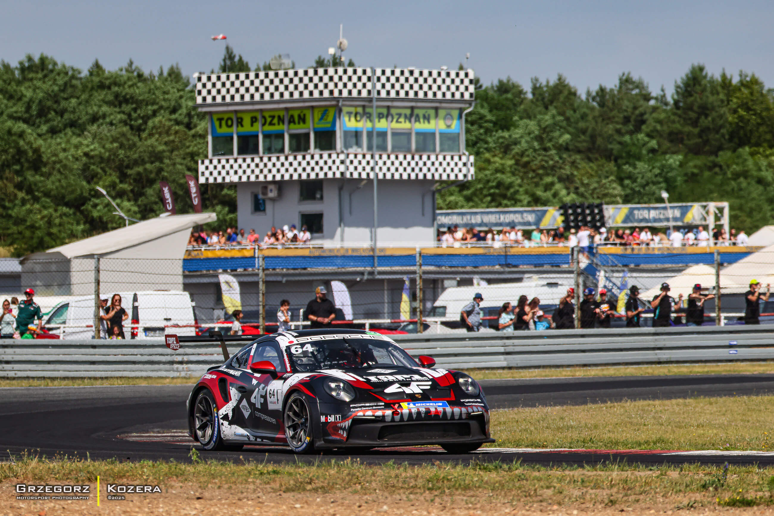 Alt Text: Samochód wyścigowy Porsche 911 GT3 Cup z numerem 64 prowadzony przez Fabiana Dybionkę pokonuje zakręt na torze Poznań, na tle budynku sędziowskiego z charakterystyczną czarno-białą szachownicą. W tle widoczni są kibice oraz fotografowie śledzący 