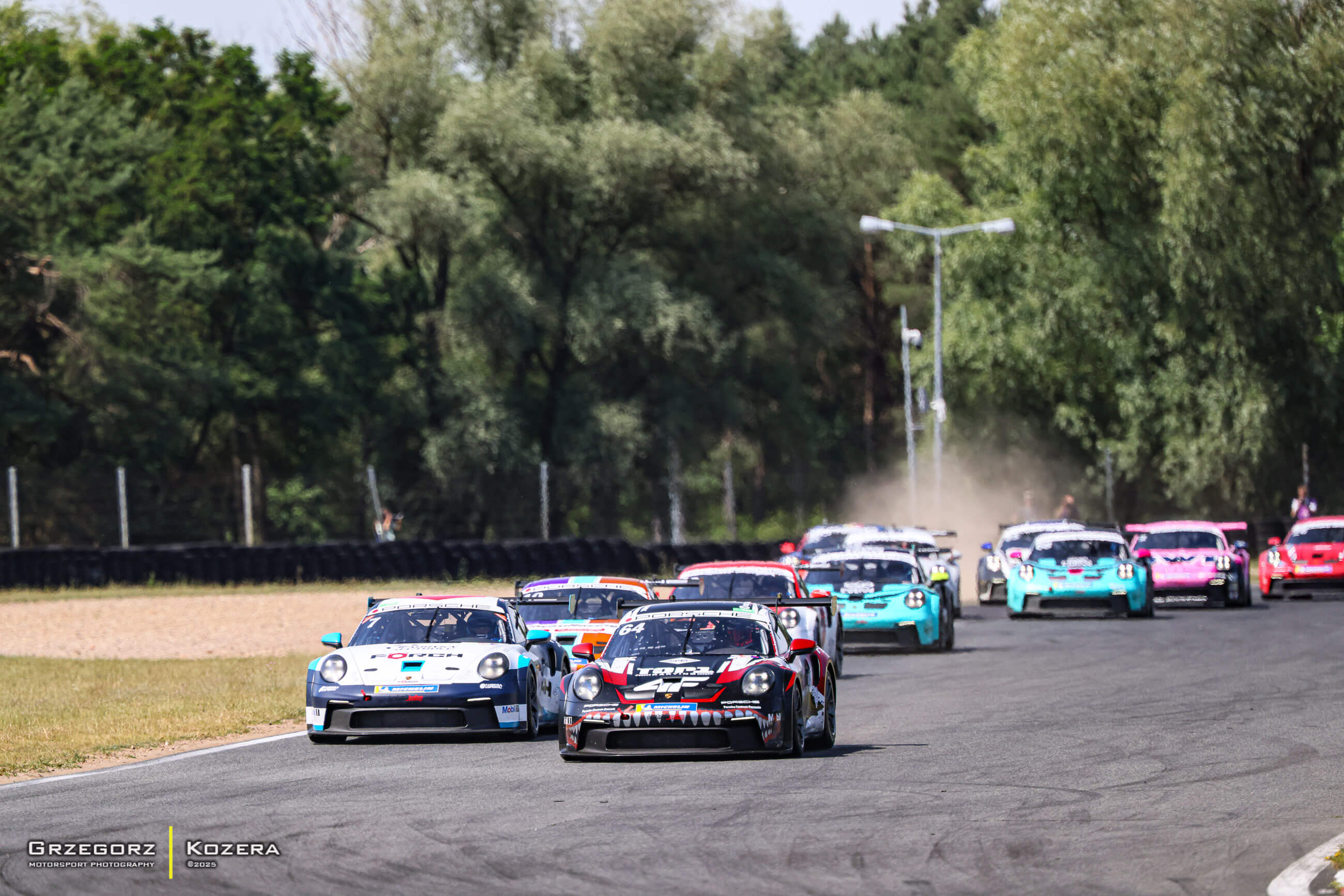 Alt Text: Samochód wyścigowy Porsche 911 GT3 Cup Fabiana Dybionki z numerem 64 i charakterystyczną grafiką rekina prowadzi stawkę na torze wyścigowym, za nim podążają inne auta serii wyścigowej. W tle widać unoszący się kurz i otaczające tor drzewa. Zdjęci